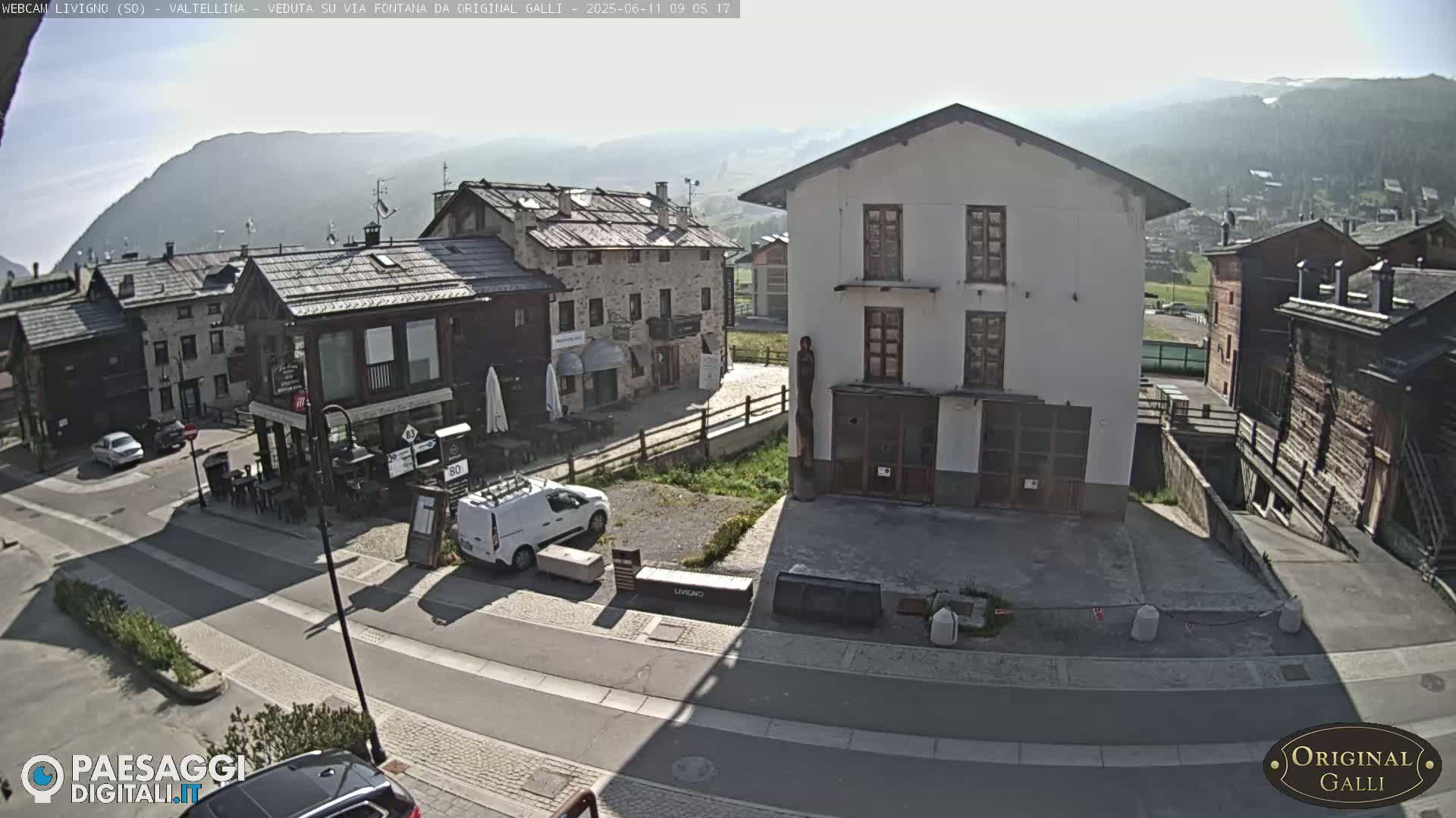 A sunny view of a village street scene nestled in a valley, surrounded by mountains.