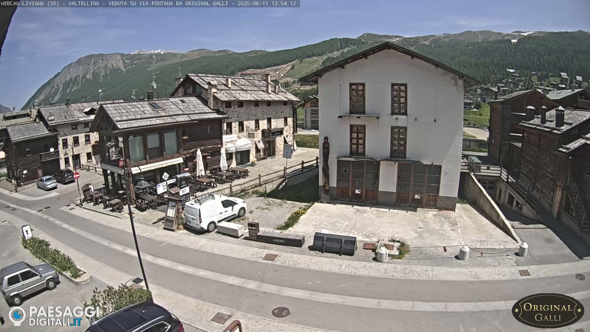 A sunny mountain village scene shows a street with parked cars, buildings of varying architectural styles, and outdoor seating areas in front of a cafe.