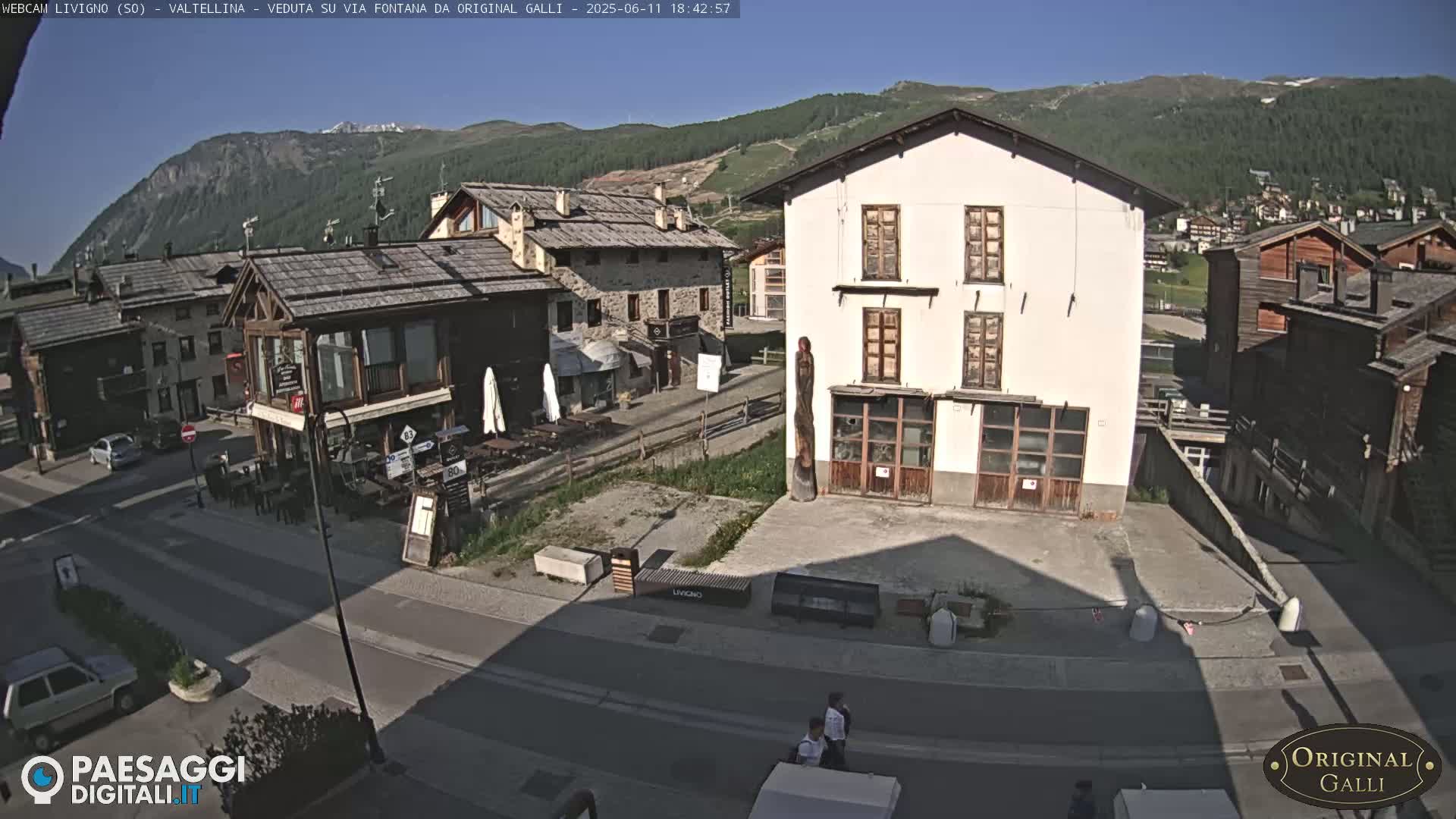 A sunny view of a village street scene in the mountains, showing several buildings, a few cars, and some pedestrians.