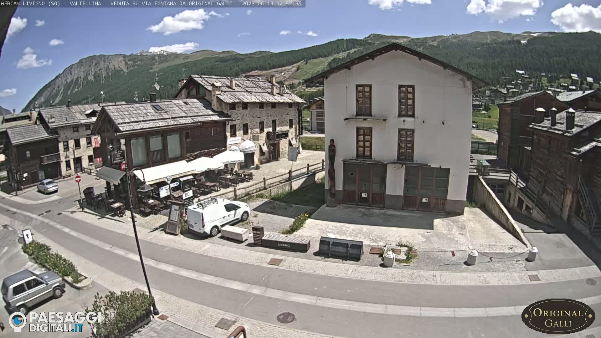 A sunny view of a village street scene with stone buildings, a few cars, and outdoor seating areas, nestled against a backdrop of mountains.