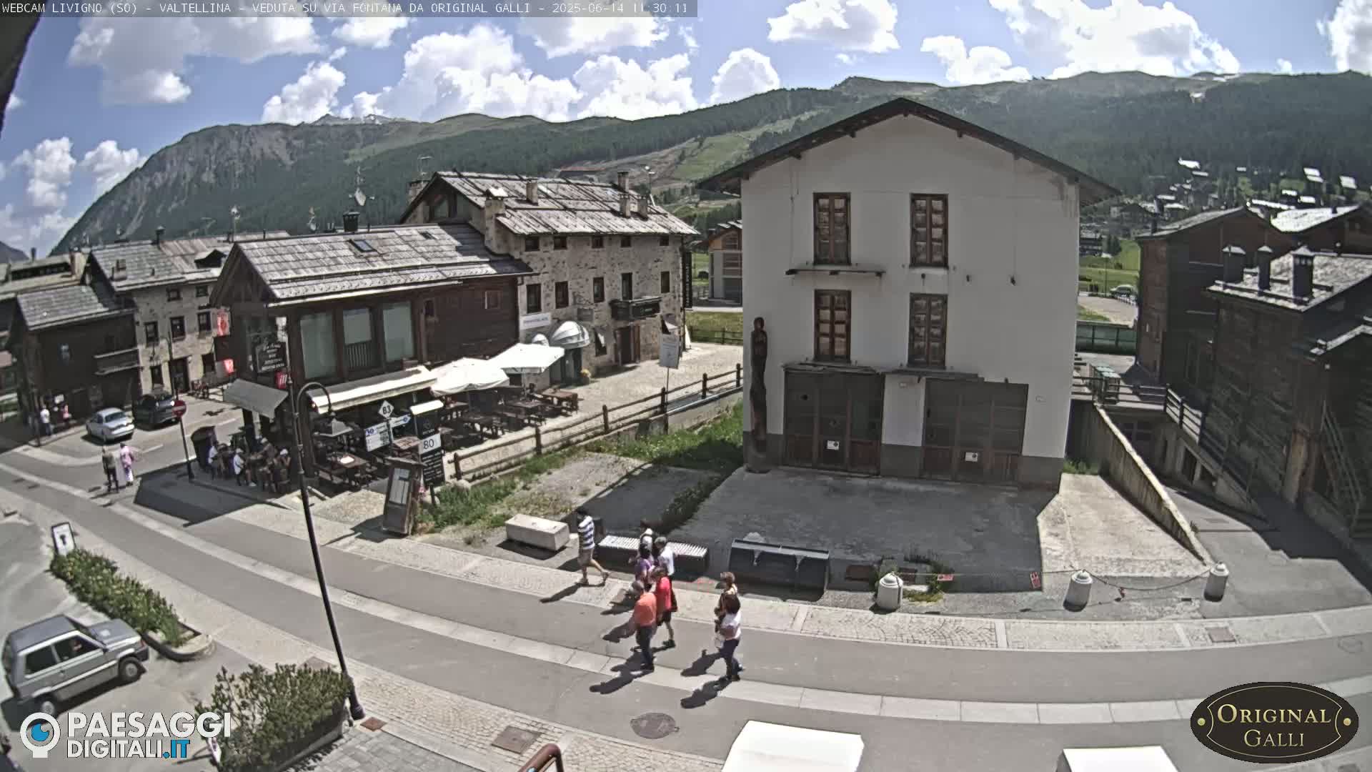 A sunny day in a mountainous village shows a town square with pedestrians, parked cars, and several buildings, including a restaurant with outdoor seating.