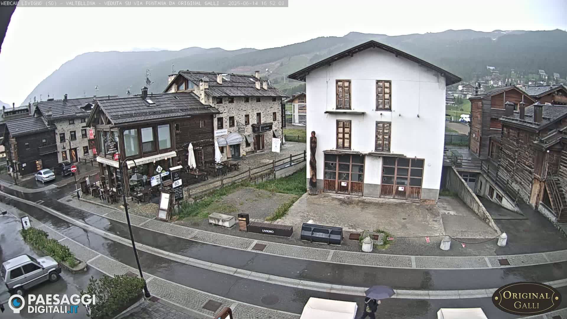 A wet, village street scene shows several buildings, some stone and some wood, under a gray sky.