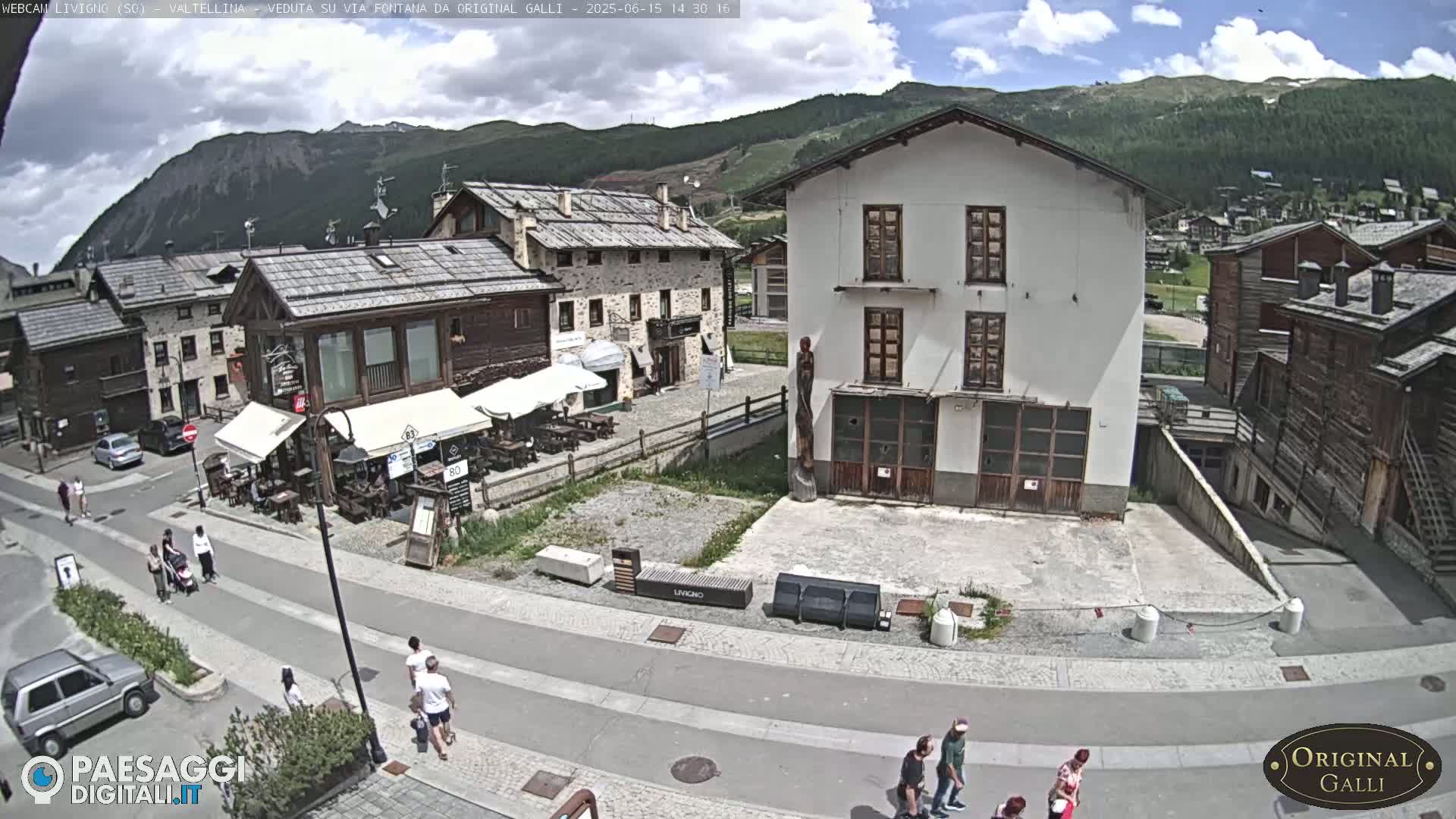 A partly cloudy day reveals a village square with pedestrians, outdoor seating at a restaurant, and buildings in a mountainous region.