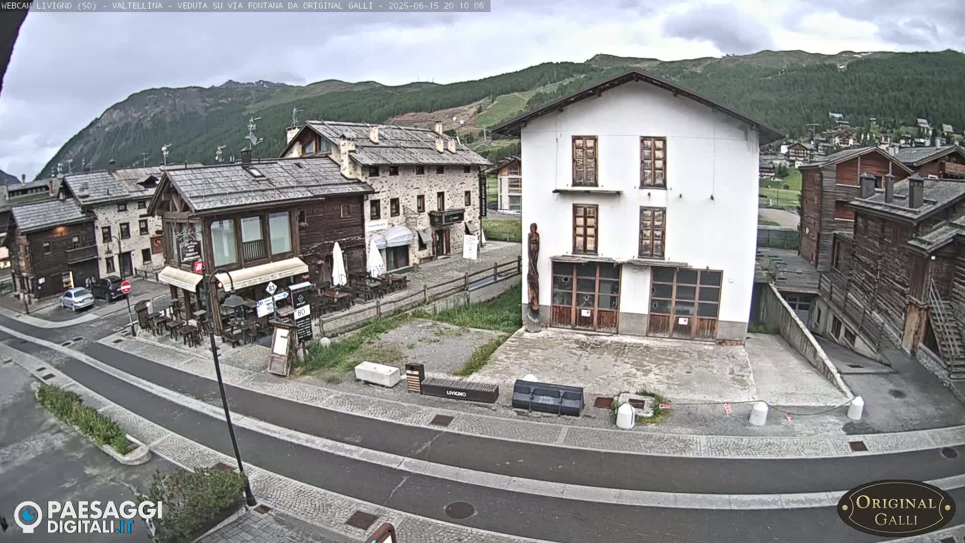 A mostly cloudy day reveals a village square with several stone buildings, some with outdoor seating, nestled against a mountainous backdrop.