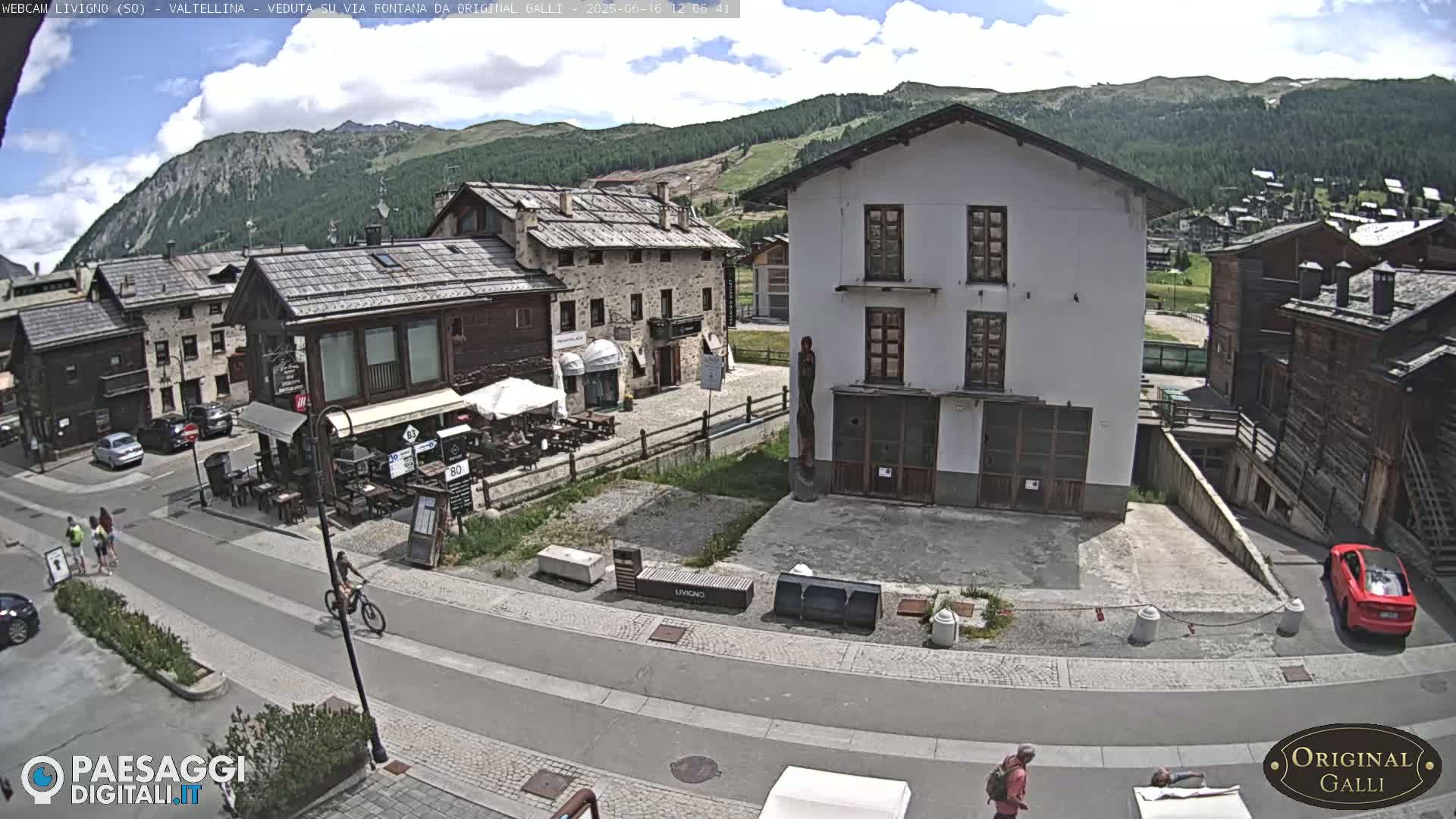 A sunny mountain village scene shows a town square with several buildings, pedestrians, and a cyclist, with mountains in the background under a partly cloudy sky.