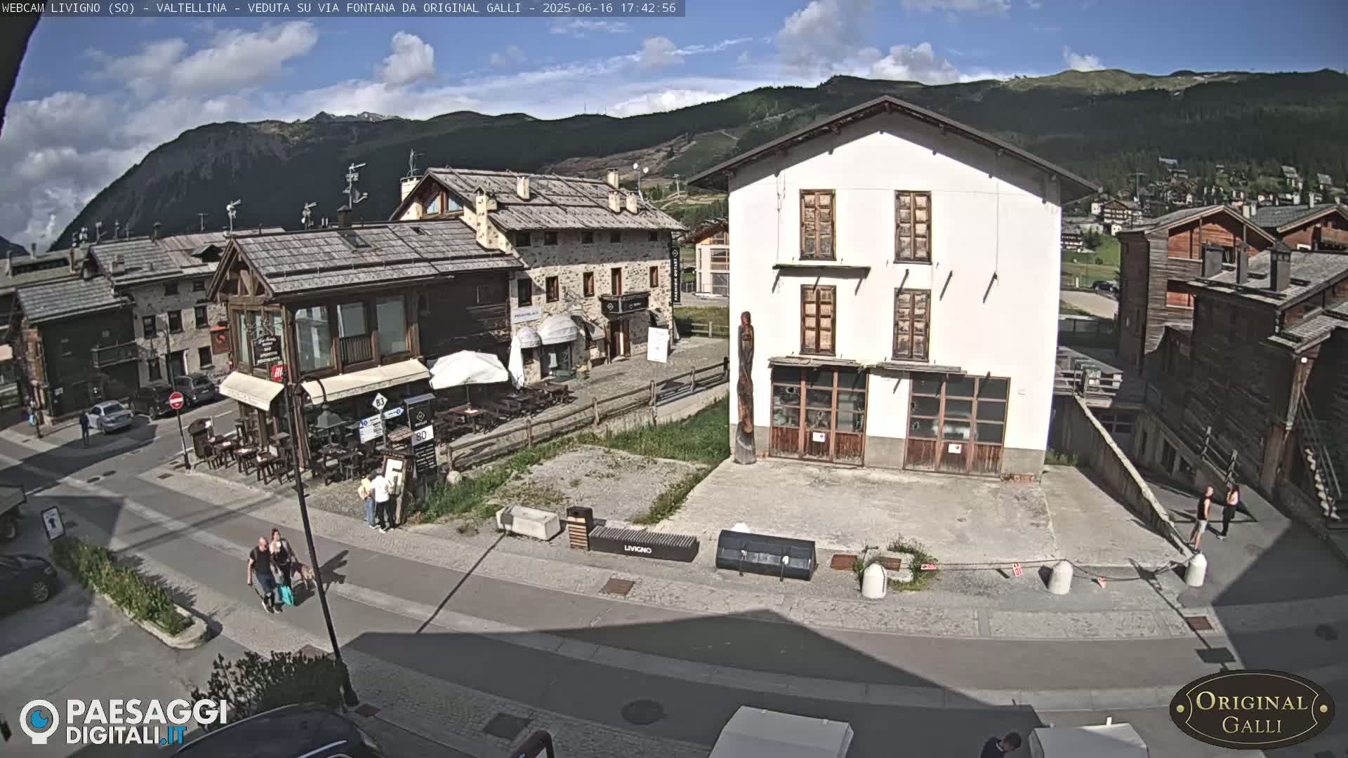 A sunny day in a village with stone and wood buildings, people walking on the street, and mountains in the background.
