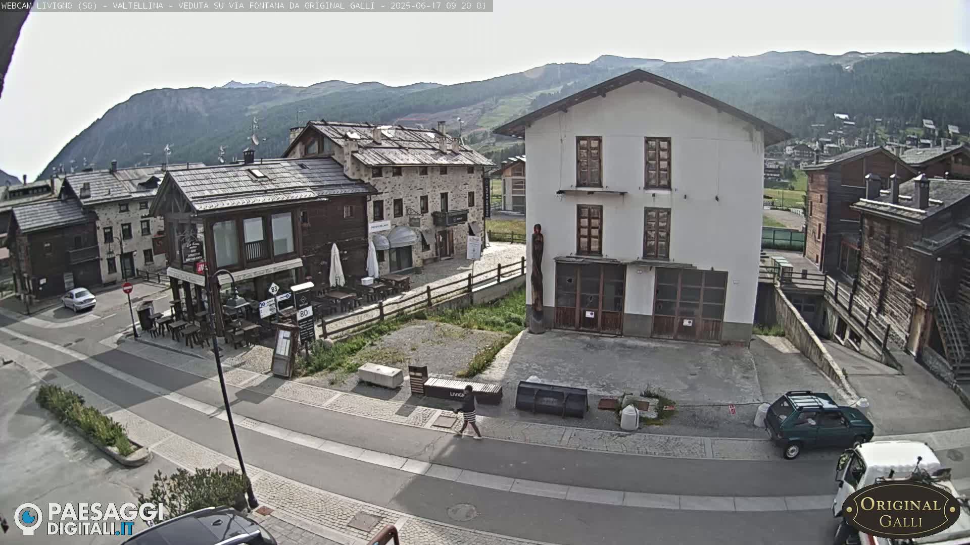 A sunny mountain village scene shows several stone and wood buildings surrounding a paved road where a person is walking and a few cars are parked.