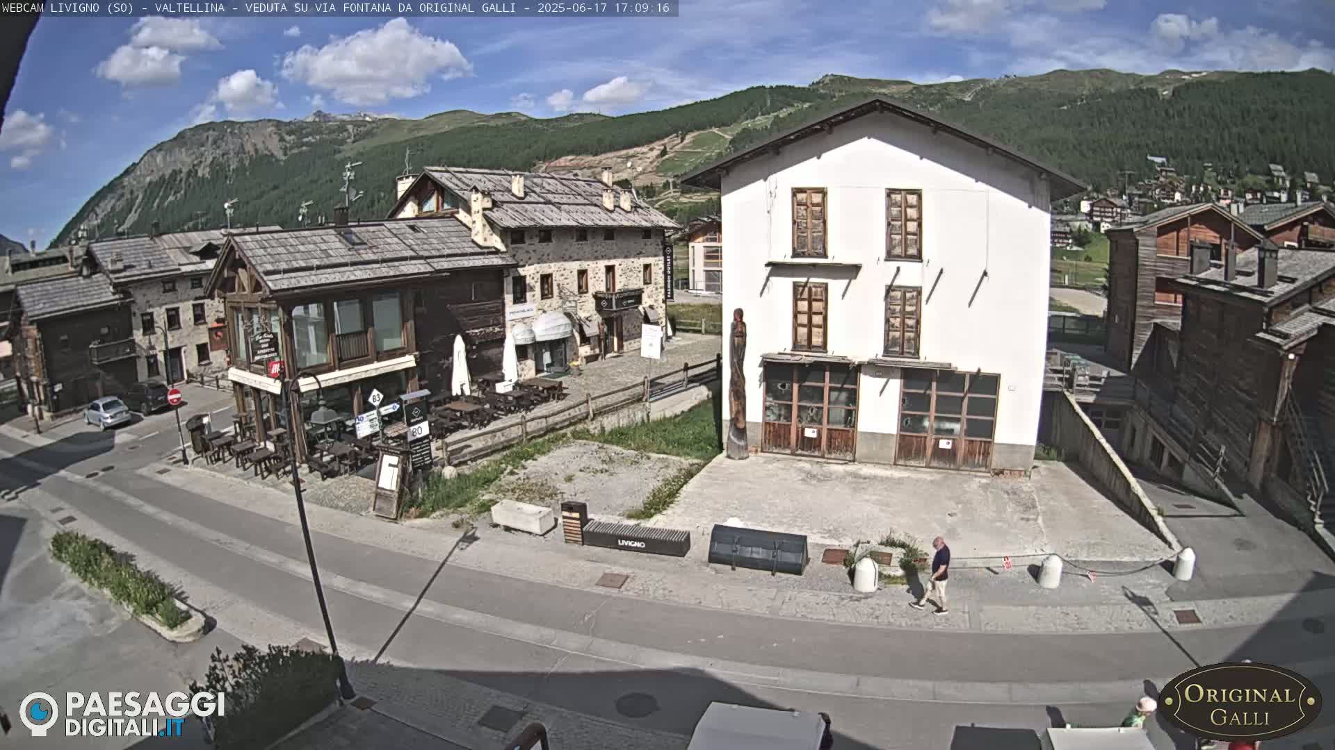 A sunny view of a town square in a mountain village, with several buildings and a few people walking.