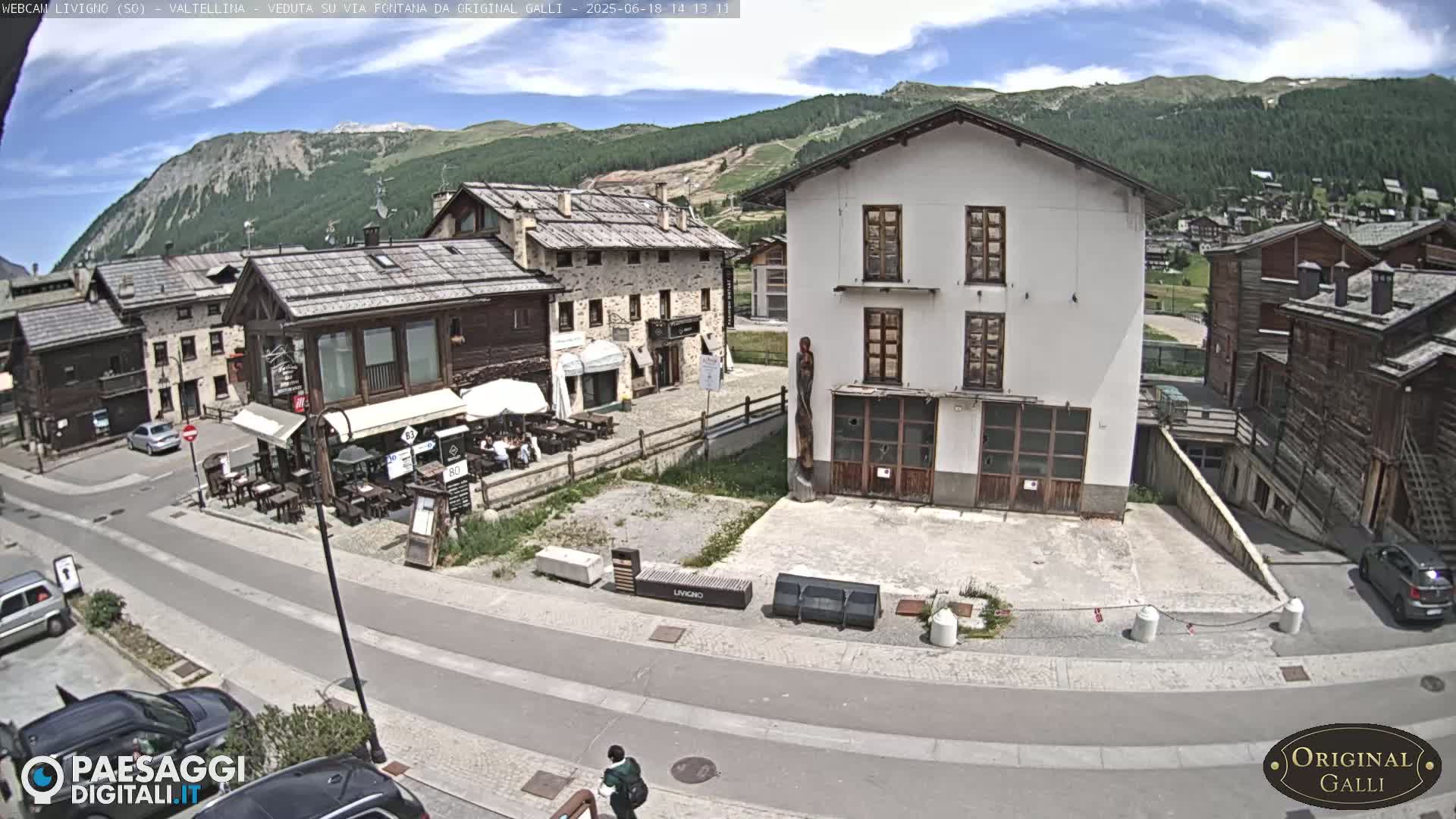 A mountain village square with several buildings, a restaurant with outdoor seating, and cars parked on a sunny day.