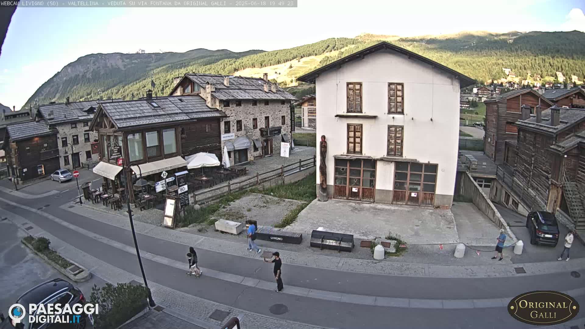 A mostly sunny mountain village street scene shows several low stone buildings, a few pedestrians, and cars parked on the side of the road.
