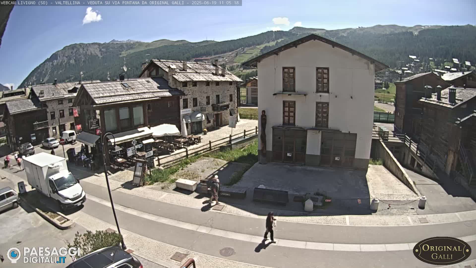 A sunny view of a village square in the mountains, with several buildings, vehicles, and a few pedestrians.