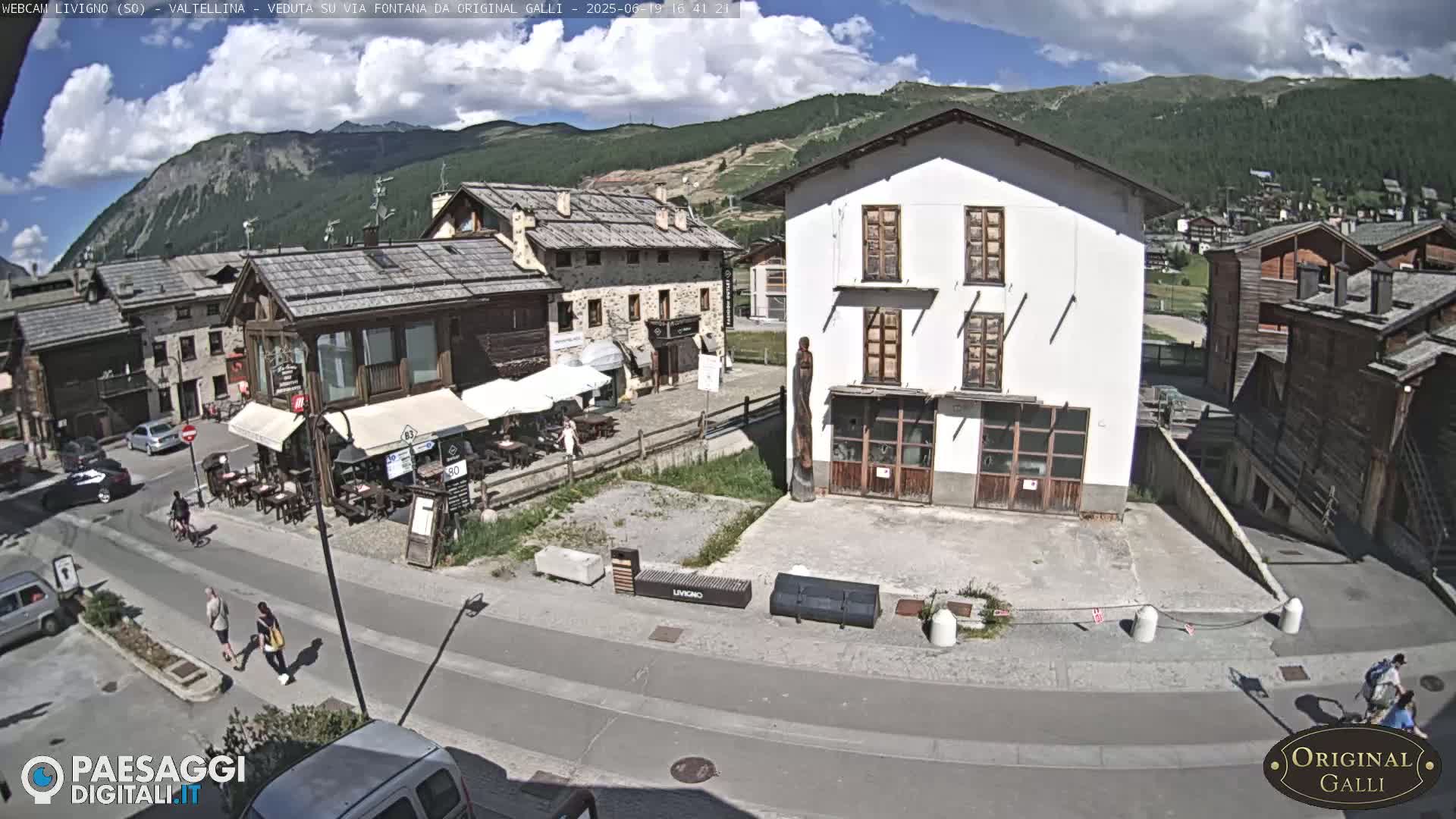 A sunny mountain village square with several buildings, a few pedestrians, and outdoor seating at a cafe.