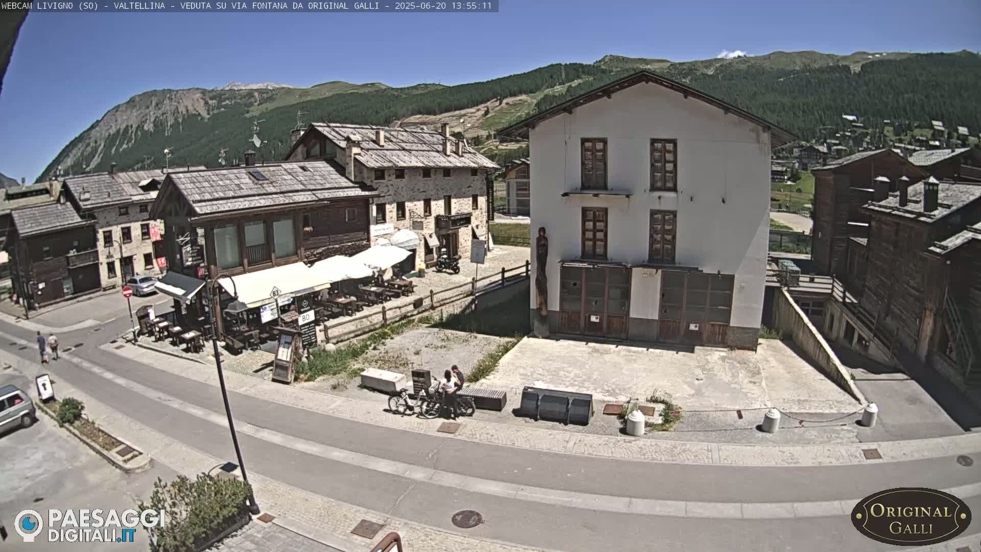 A sunny view of a village square with several buildings, outdoor seating, and a few people.