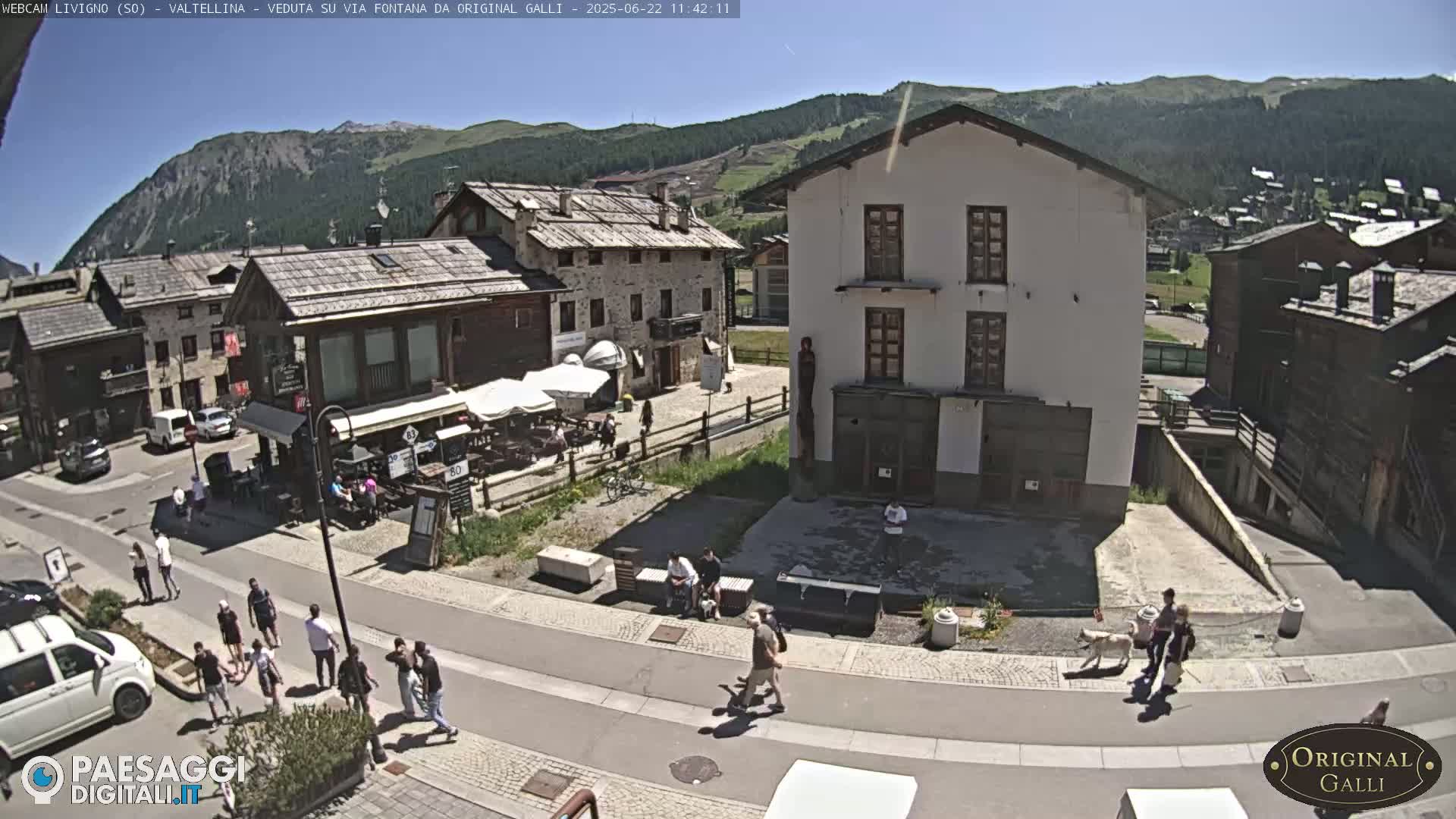 A sunny mountain village scene shows people walking on a street in front of stone buildings, with a few cars parked nearby.