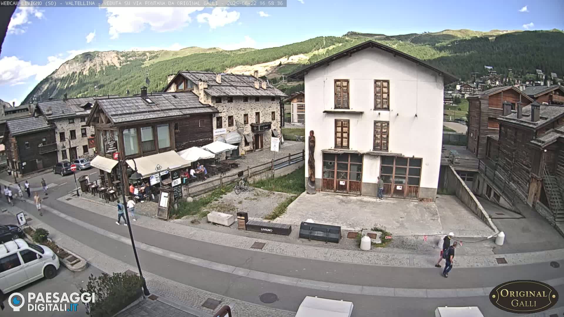 A sunny day in a mountain village with stone buildings and people walking along a paved street, near outdoor seating and a few parked cars.