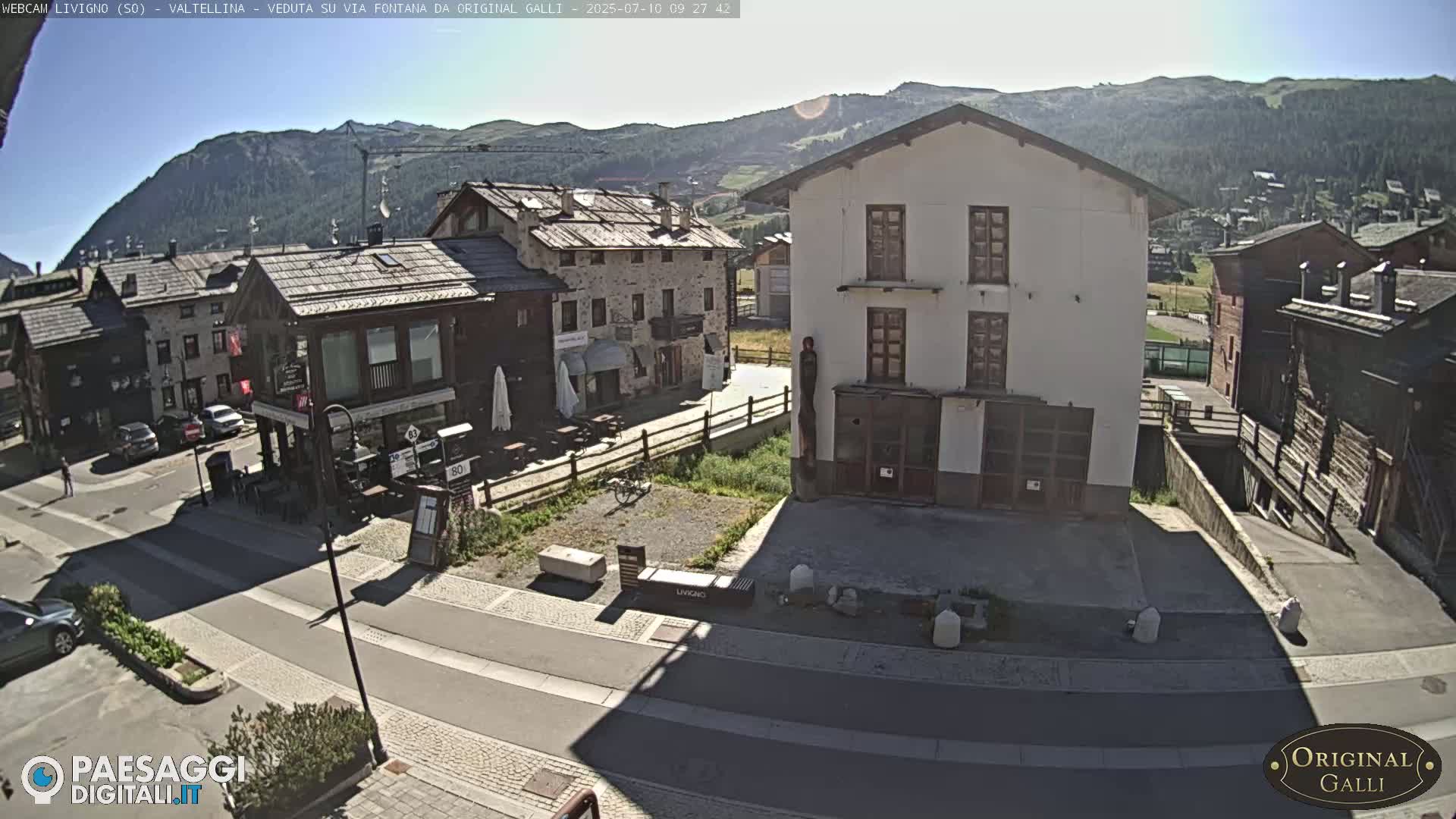 A sunny view of a village nestled in a mountain valley shows several stone and stucco buildings along a paved road.