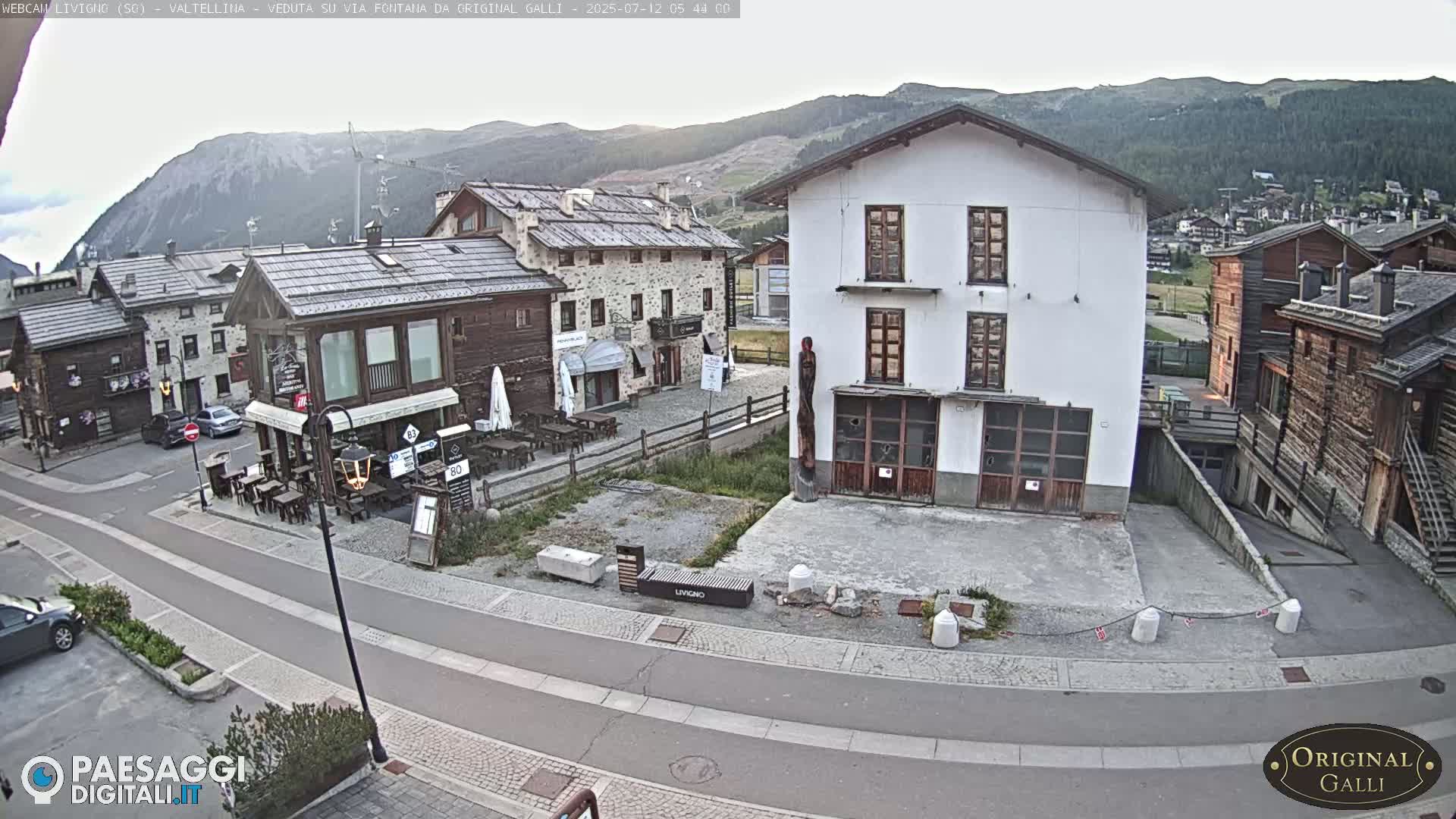 A sunny view of a village nestled in a mountain valley shows several stone and stucco buildings along a paved road.