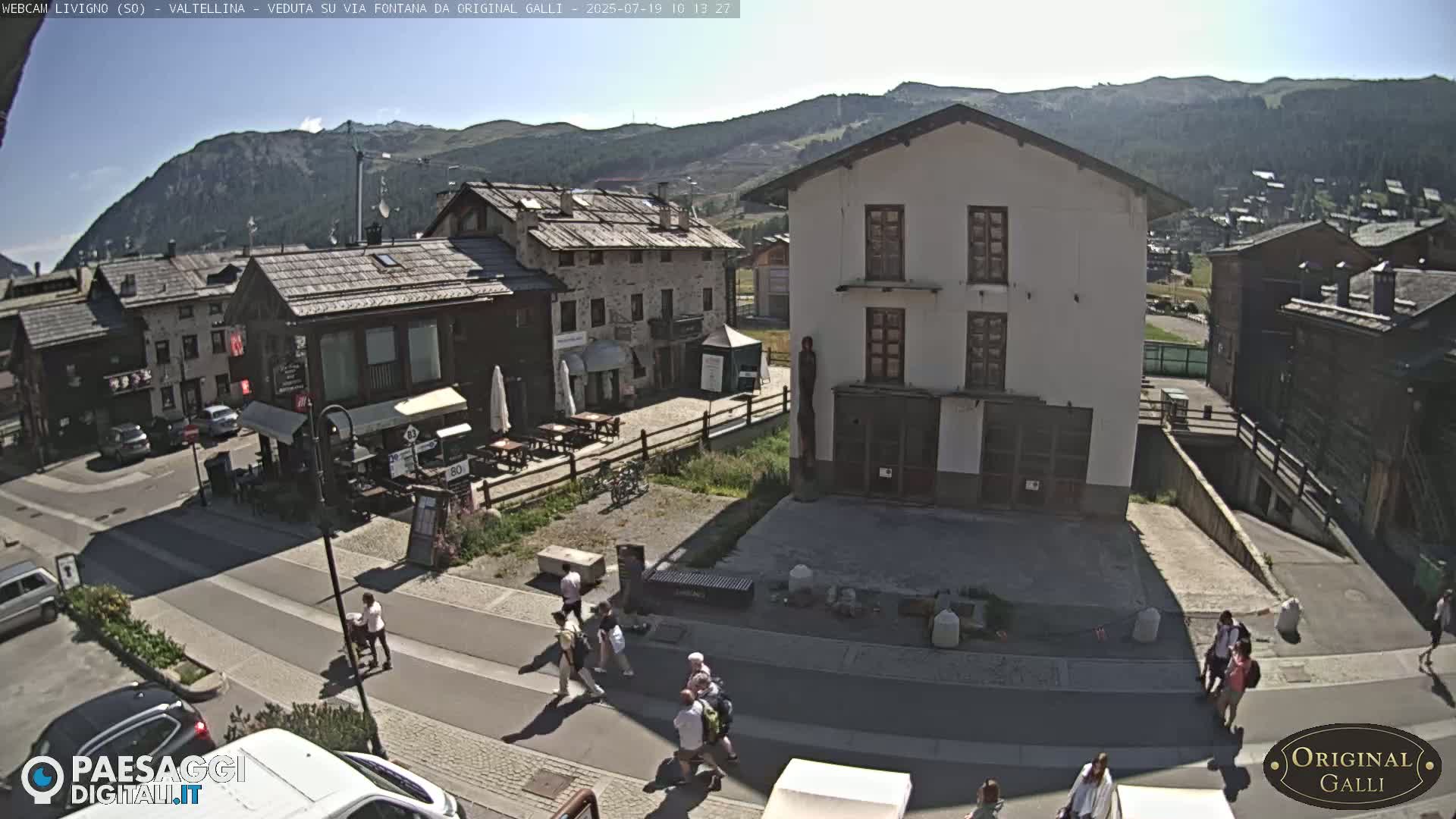 A sunny mountain village square with several buildings, pedestrians, and parked cars.