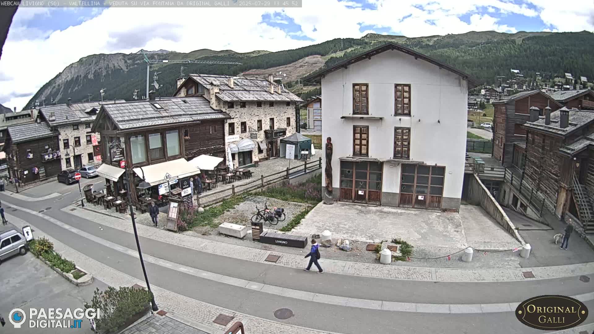 A sunny mountain village square with several buildings, pedestrians, and parked cars.
