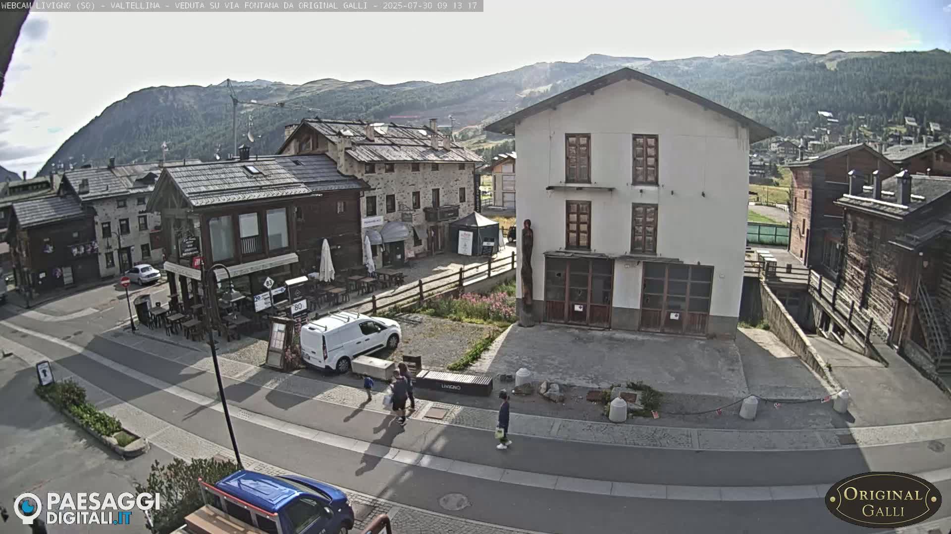A sunny mountain village square with several buildings, a few pedestrians, and parked vehicles.