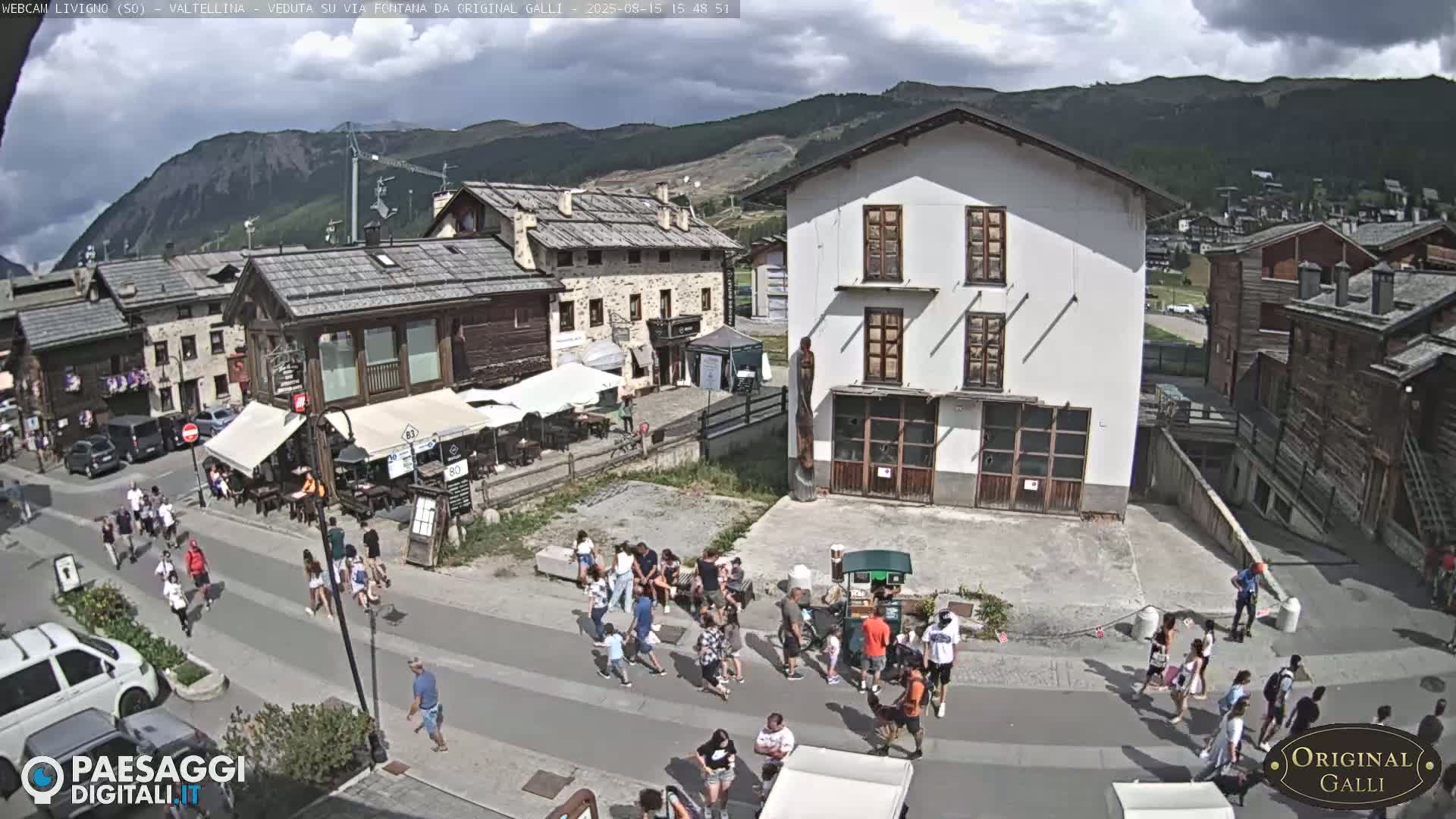 A sunny day in a village nestled in a mountain valley shows pedestrians walking along a street and sitting at outdoor cafes.