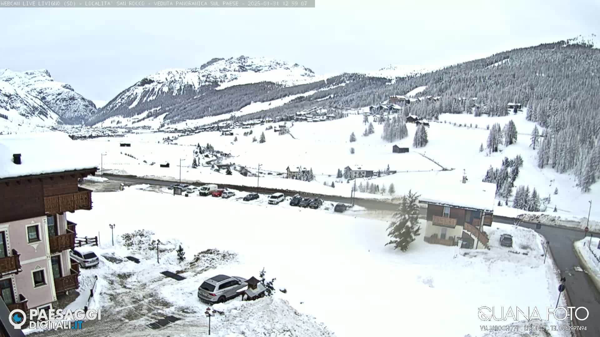 A snow-covered mountain village scene, viewed from a slightly elevated position, showing buildings, cars parked in snow, and snow-covered trees and mountains in the background under an overcast sky.