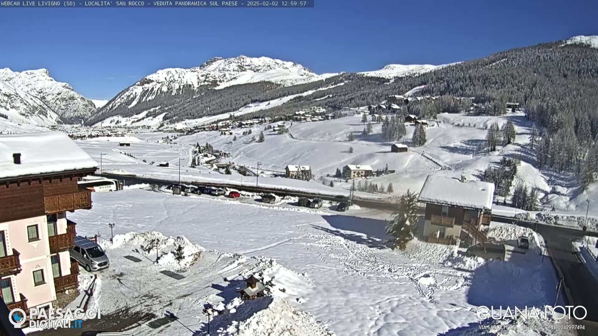 A snow-covered mountain village is seen on a sunny day, with cars parked near several buildings and a road running through the valley.