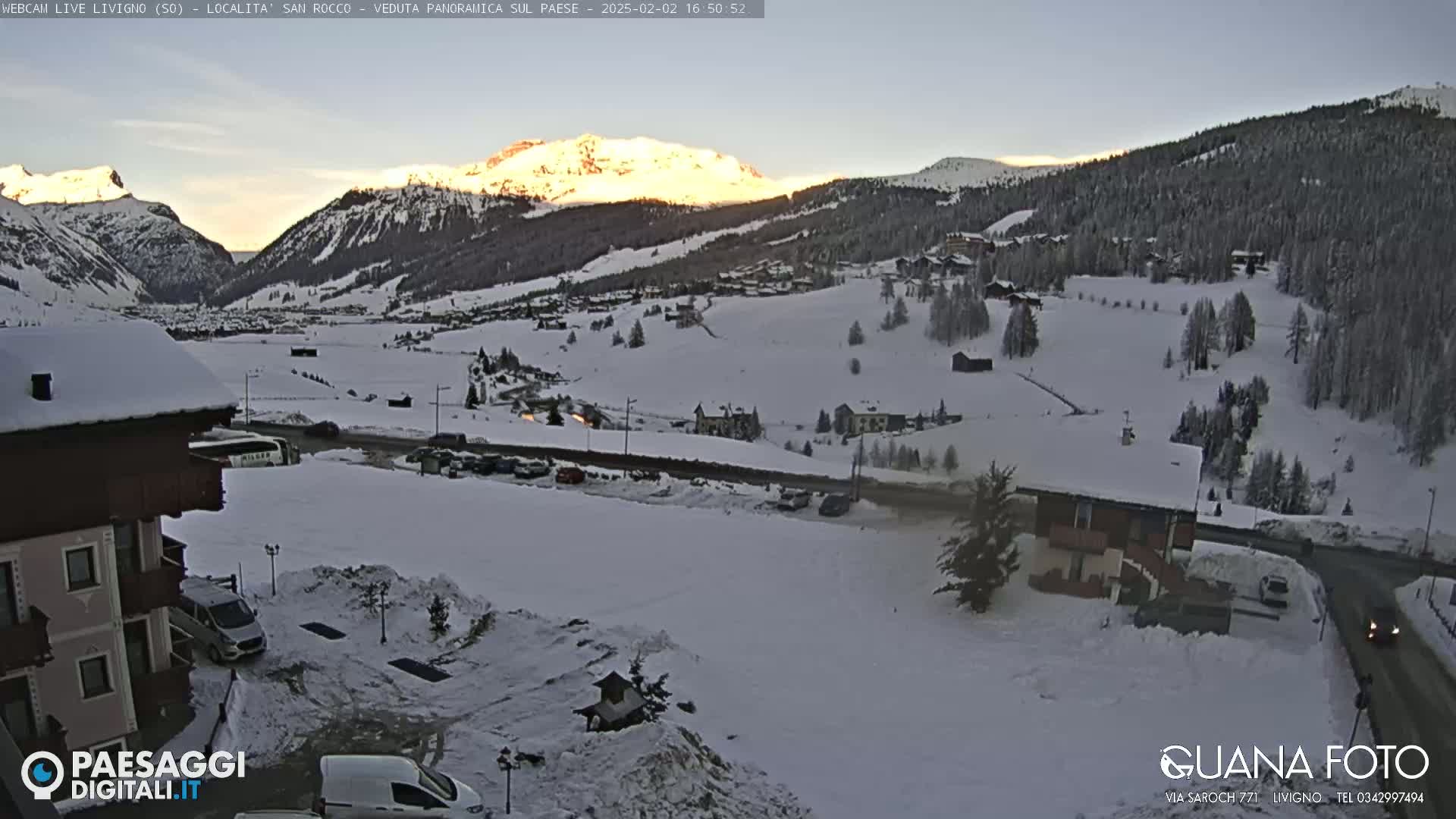 A snow-covered mountain village at dusk, with a sunlit mountain range in the background.