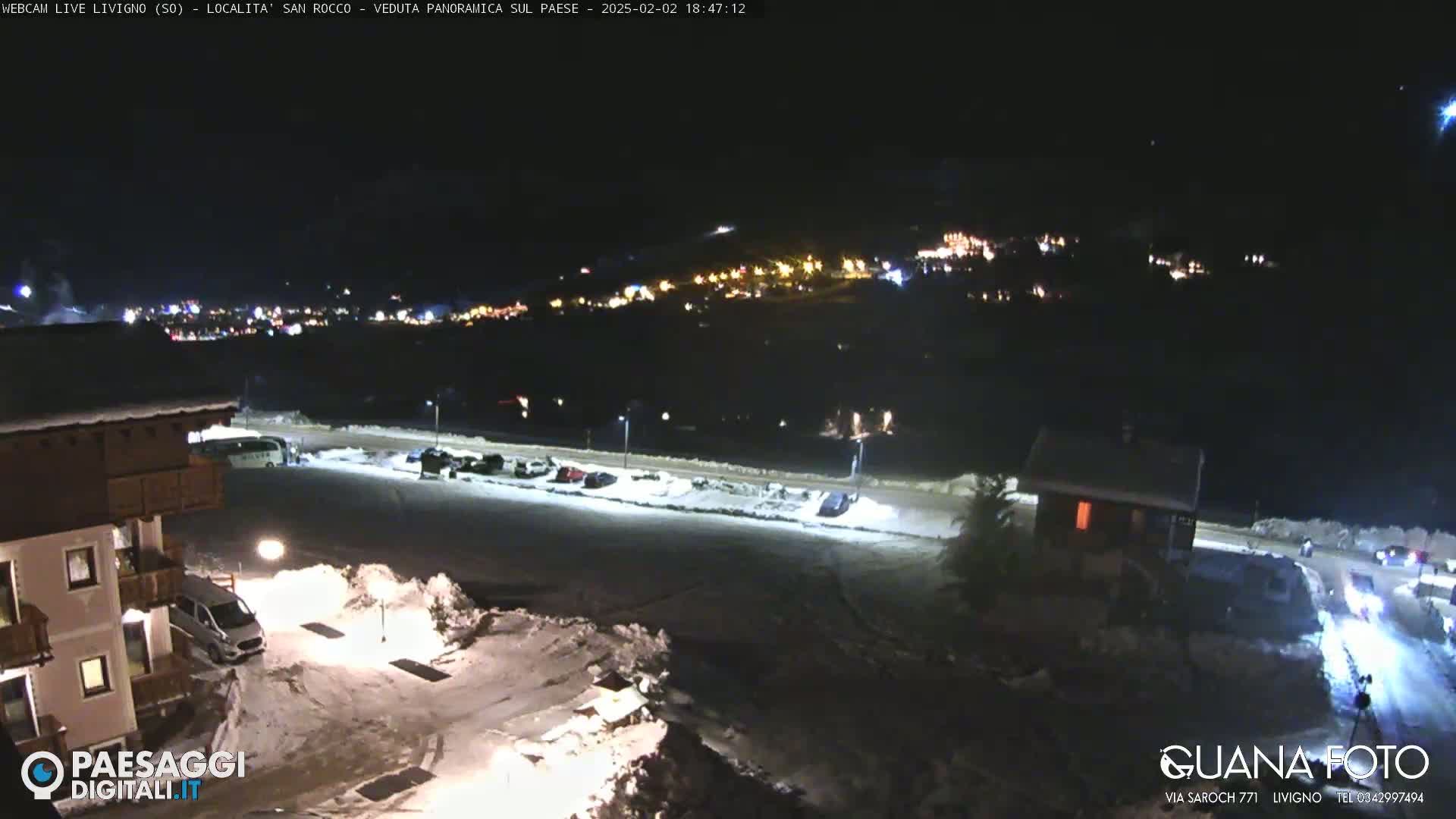A nighttime, snowy scene shows a village in the distance illuminated by lights, with a parking lot and buildings in the foreground under artificial lighting.
