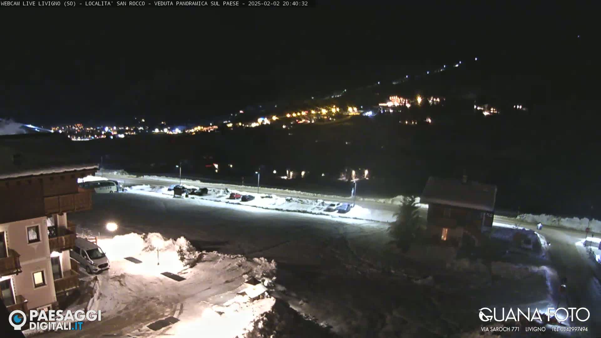 A nighttime, snow-covered parking lot and buildings are illuminated, with a town's lights visible on a hillside in the distance.