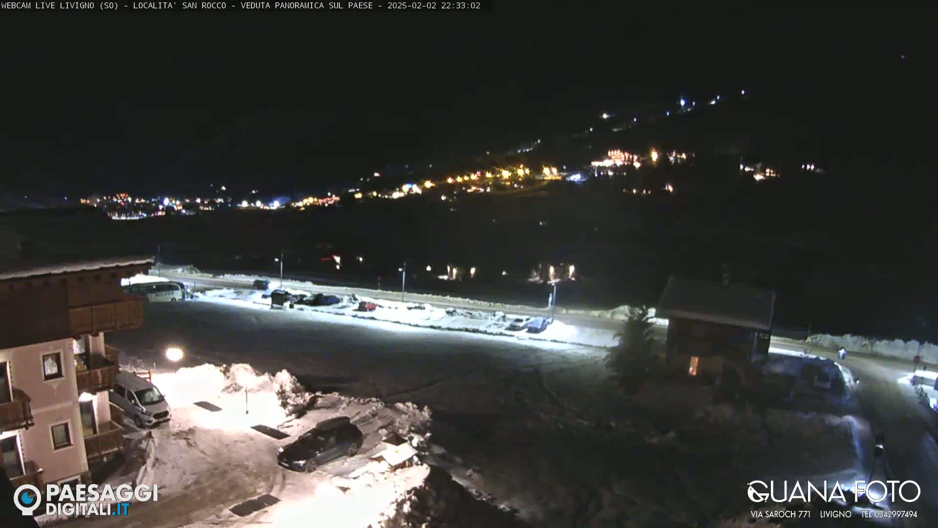 A nighttime, snow-covered parking lot and surrounding buildings are illuminated, with a distant town showing lights on a hillside in the background.