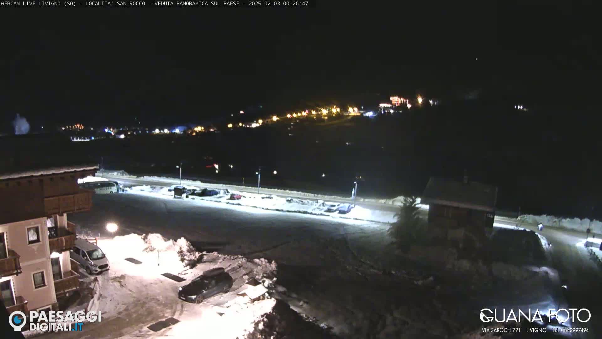A nighttime, snow-covered parking lot and village are seen under a dark sky, with lights illuminating buildings in the distance.