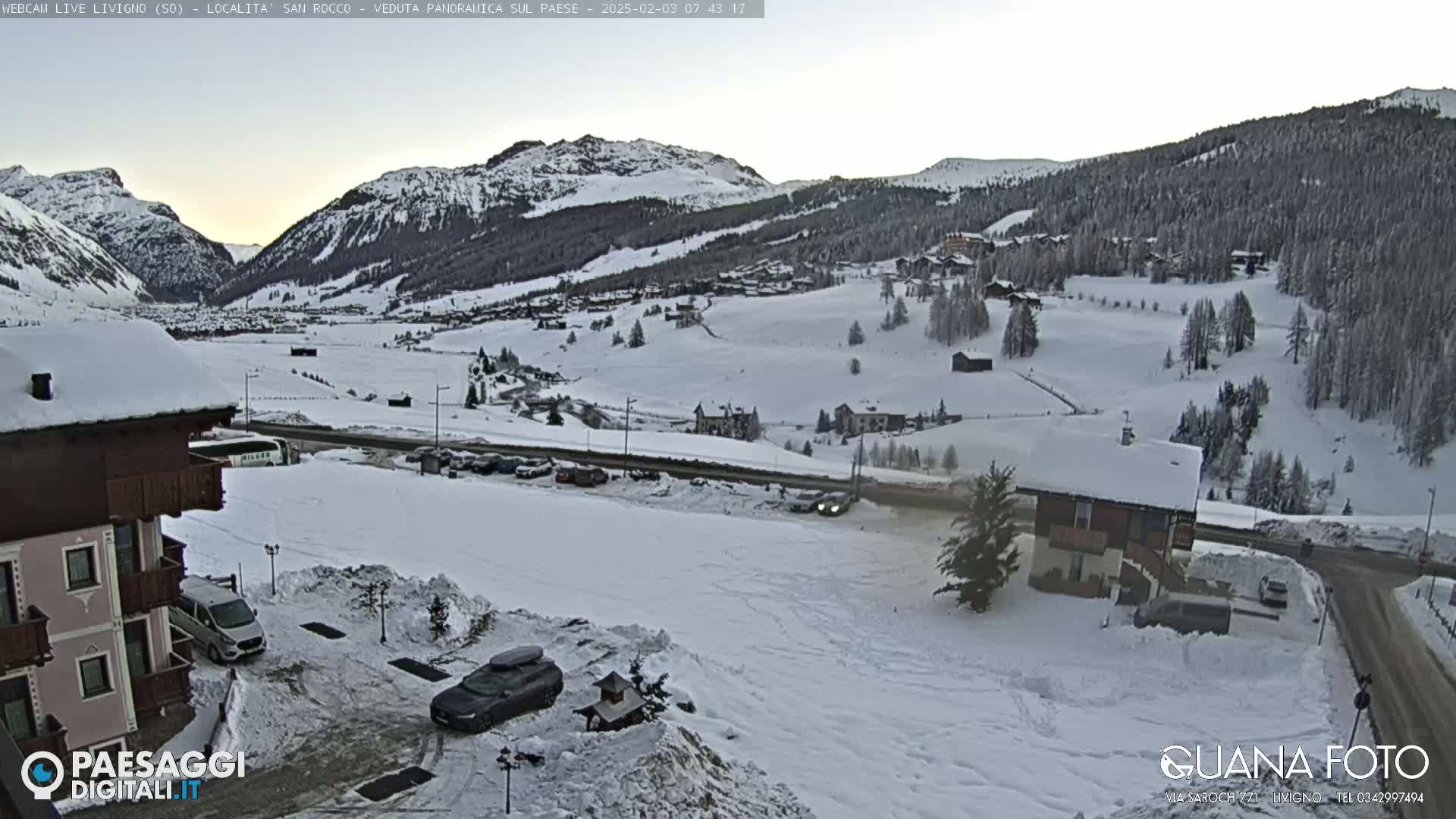 A snow-covered mountain valley scene at dawn, with buildings, cars, and a road visible.