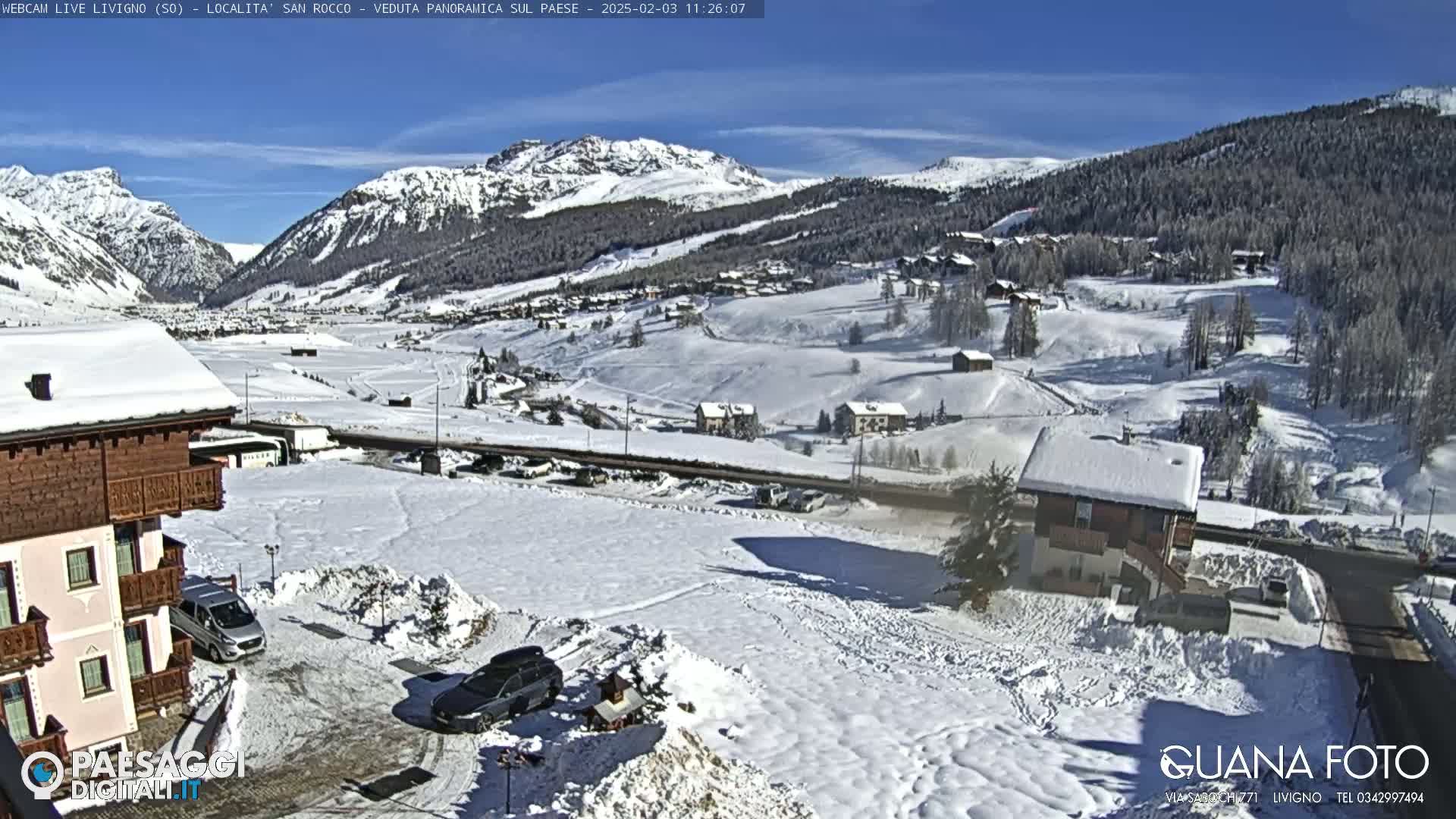 A snow-covered mountain village is seen on a sunny day, with several buildings visible and cars parked in the snow.