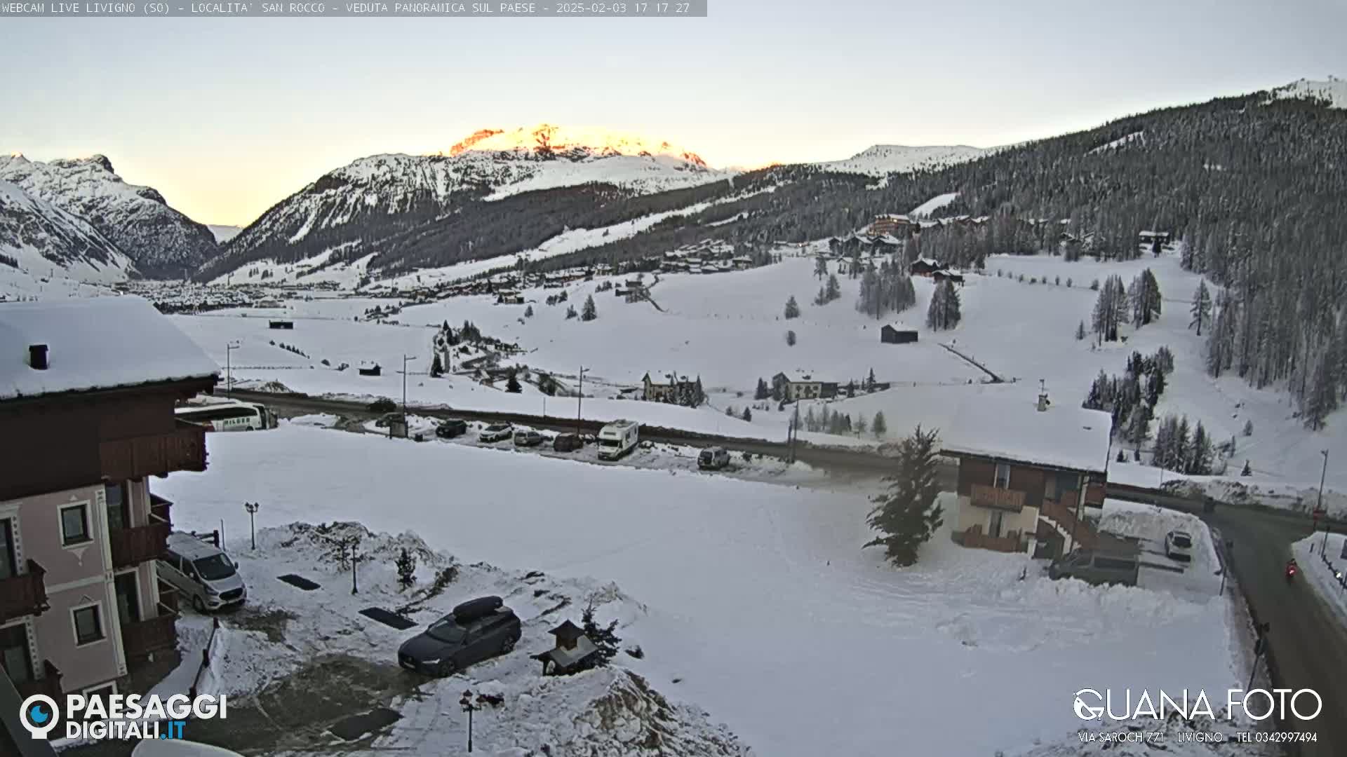 A snow-covered mountain valley at sunset, with a village nestled amongst the slopes and several cars parked in a lot near a building.