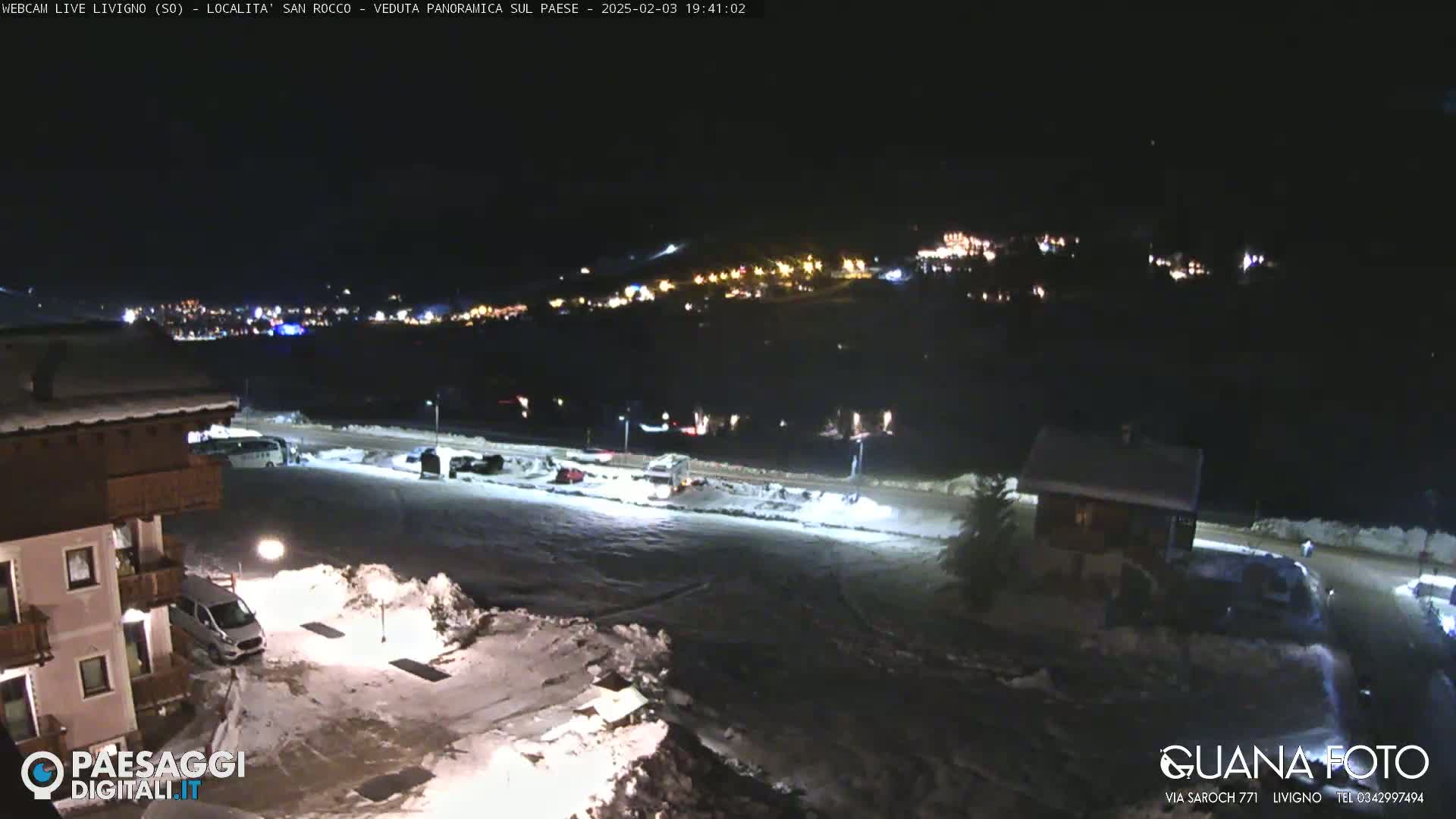A nighttime, snow-covered parking lot and residential area are illuminated, with a distant town twinkling with lights in the background.
