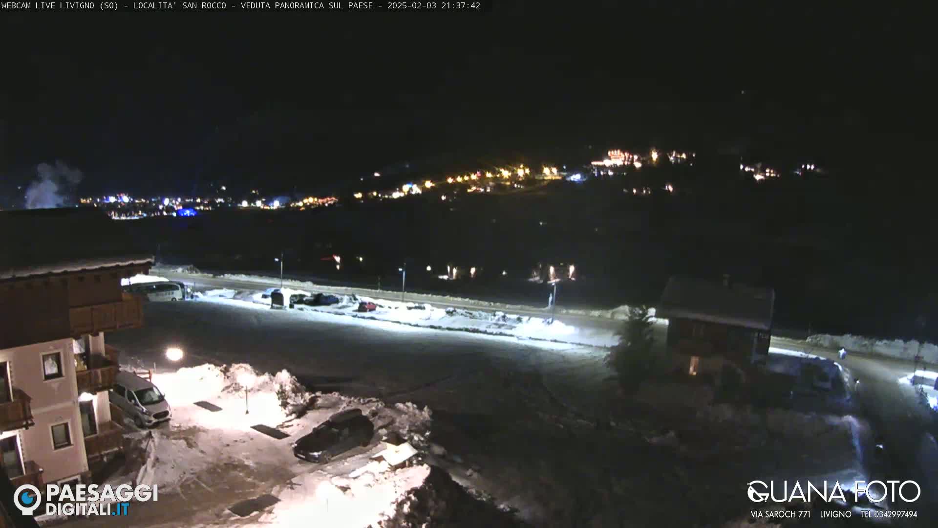 A nighttime, snowy scene shows a town in the distance illuminated by lights, with a parking lot and buildings in the foreground under artificial light.