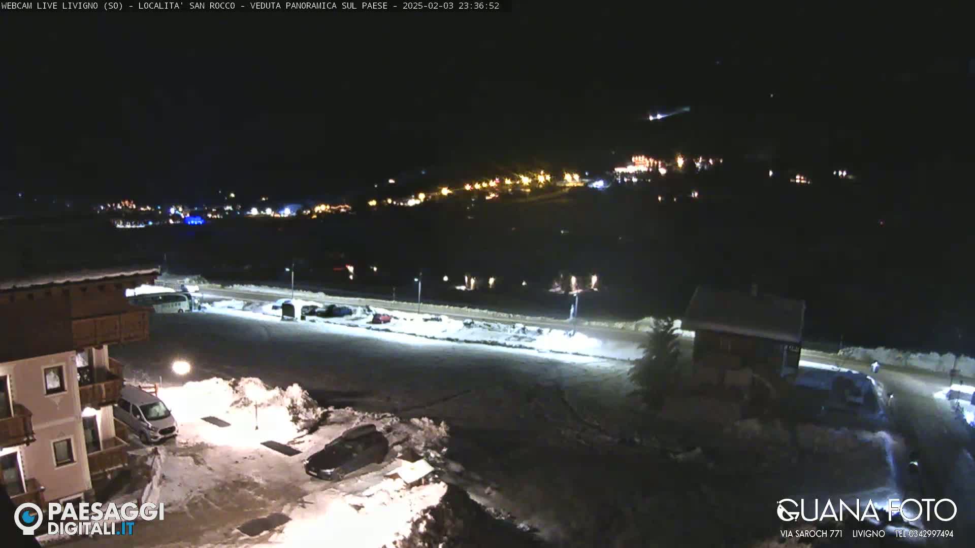 A nighttime, snow-covered parking lot and buildings are illuminated, with a town's lights visible in the distance.