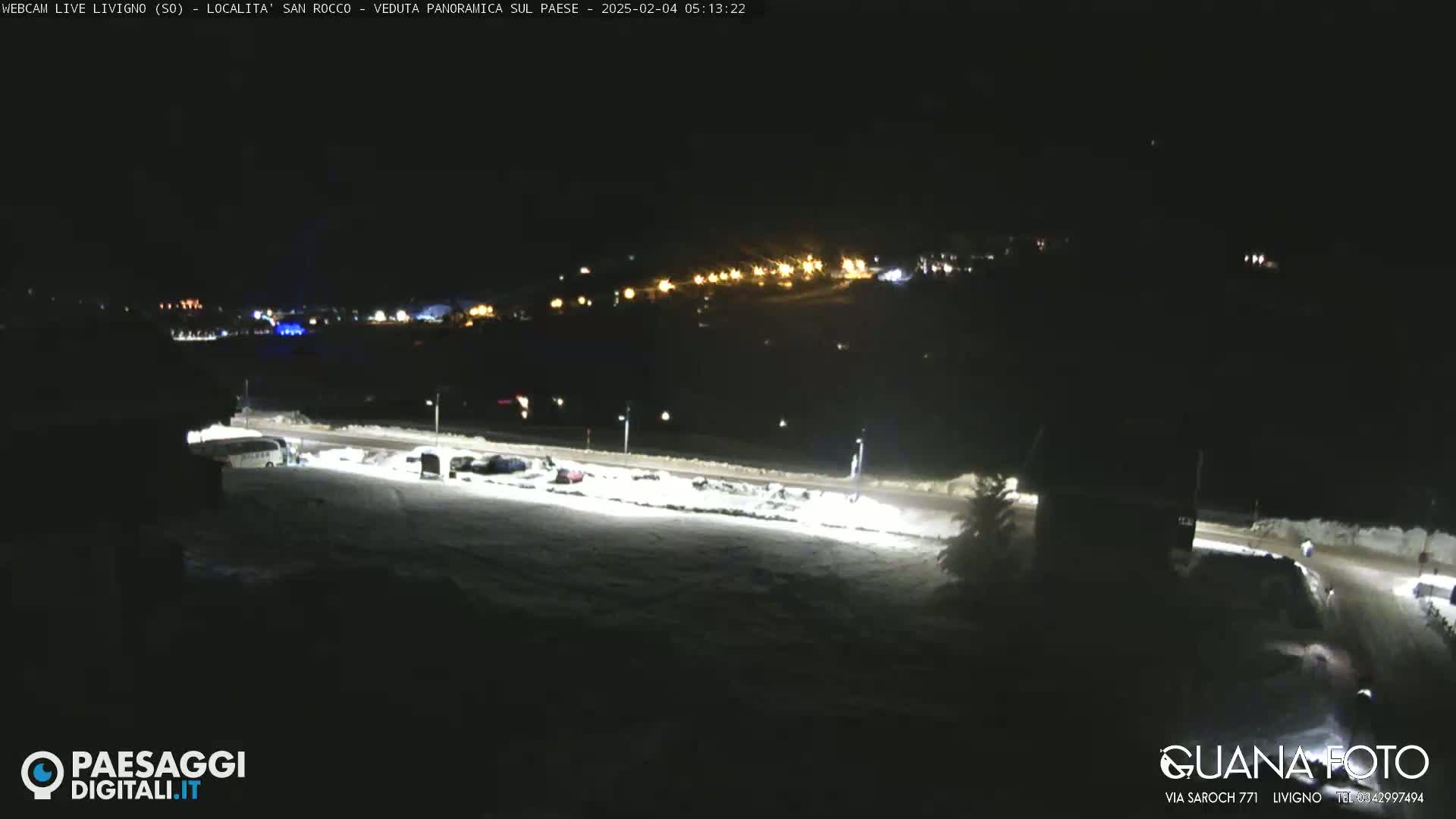A nighttime, snow-covered parking lot and road are illuminated, with a distant town showing lights in the background.