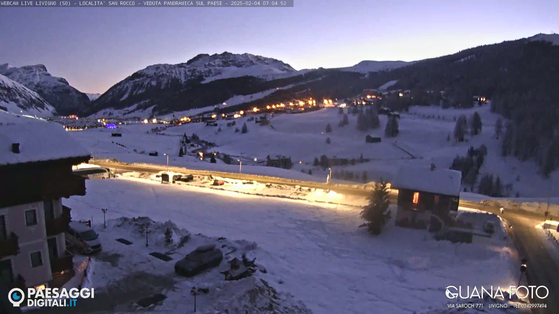 A snow-covered mountain village is illuminated at twilight, with a road and parking areas visible in the foreground.