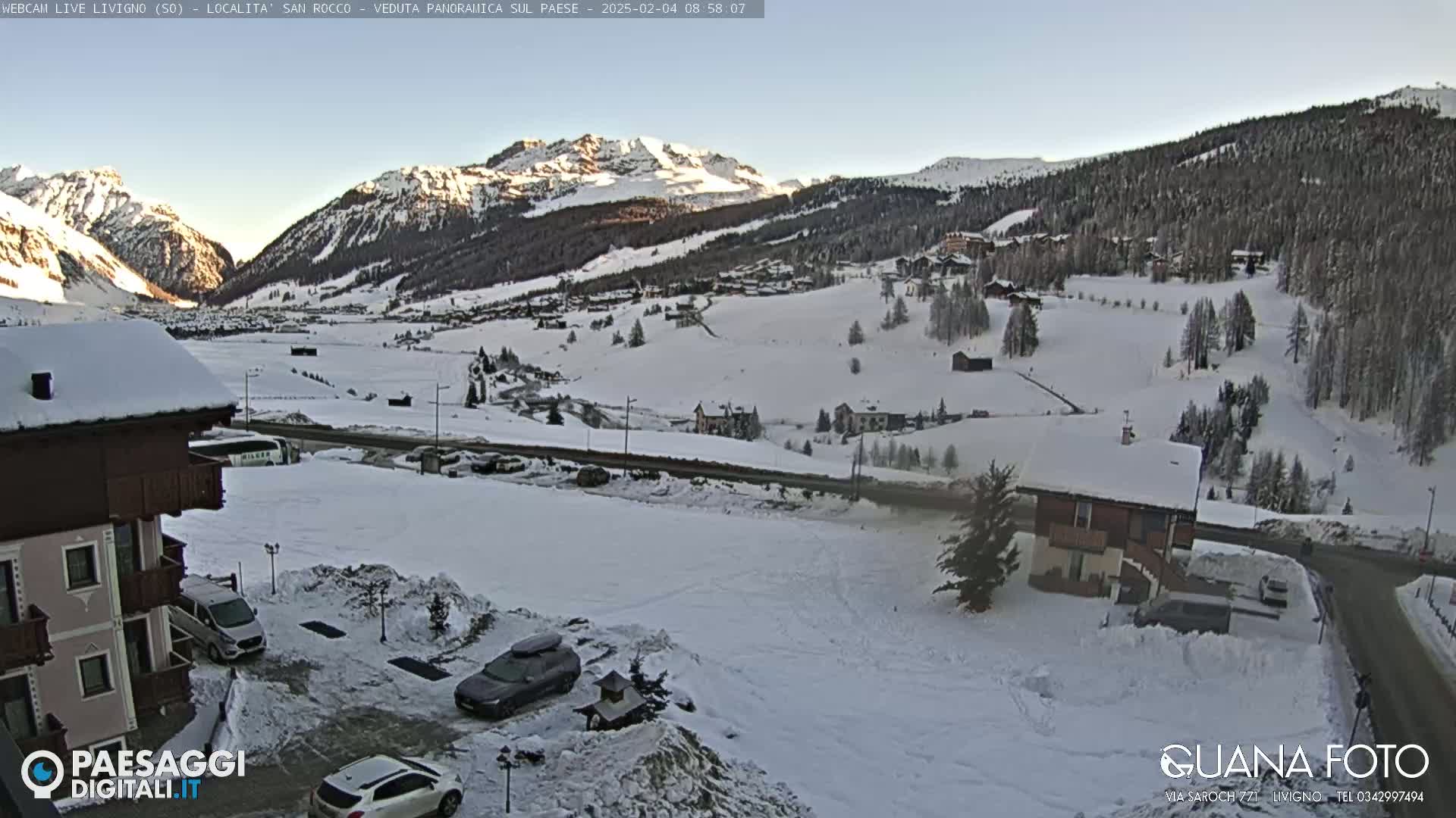 A snow-covered mountain valley scene at sunrise, showing buildings, cars, and a road, with a clear sky.