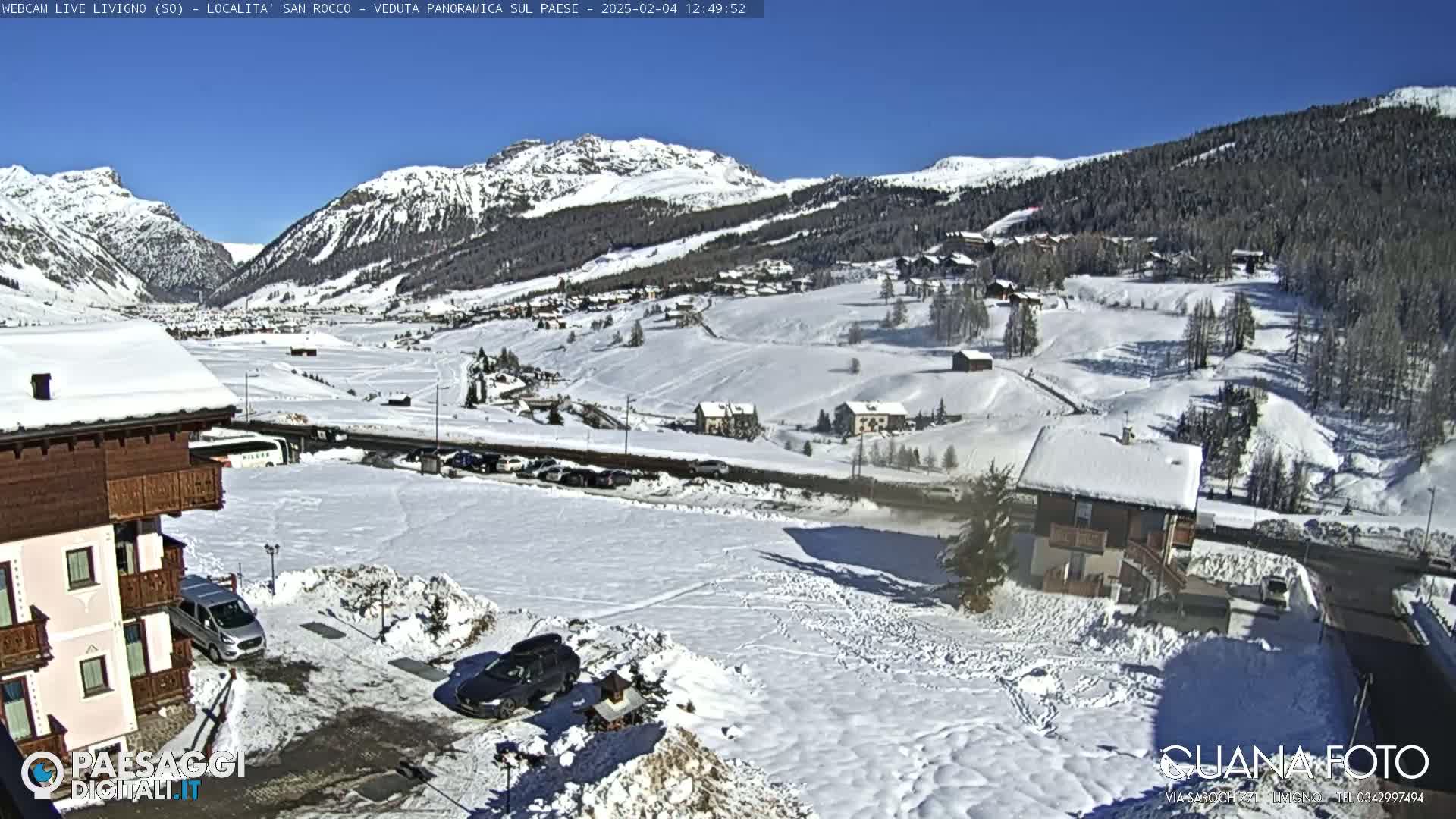 A snow-covered mountain village scene under a clear blue sky, with several buildings and cars visible.