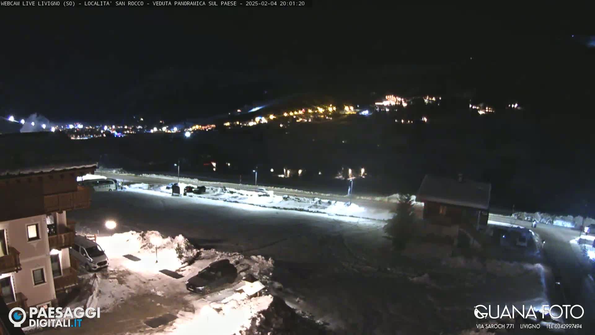A nighttime, snow-covered parking lot and small buildings are shown, with a distant town illuminated in the background under a dark sky.