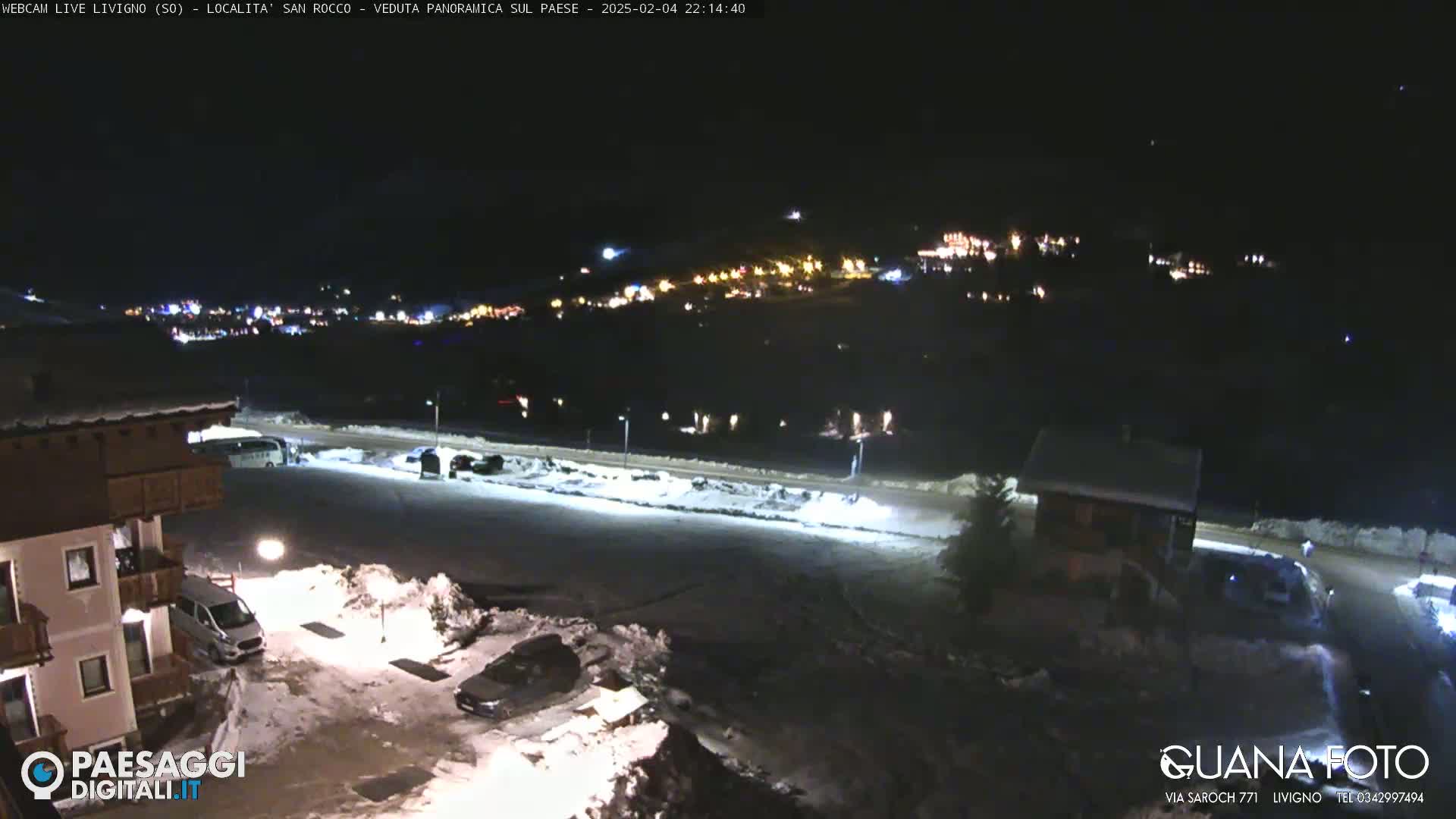 A nighttime, snow-covered parking lot with buildings and a distant, illuminated town in the background.
