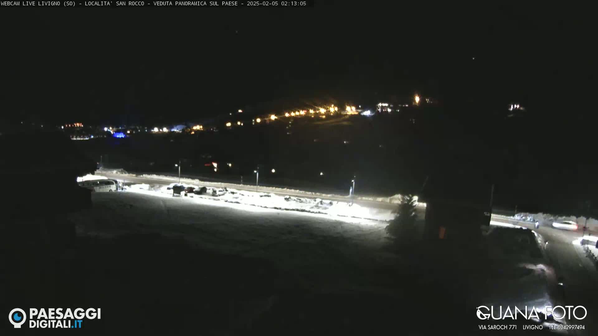 A nighttime, snow-covered parking lot with a few vehicles is illuminated, overlooking a distant village with lights under a clear, dark sky.