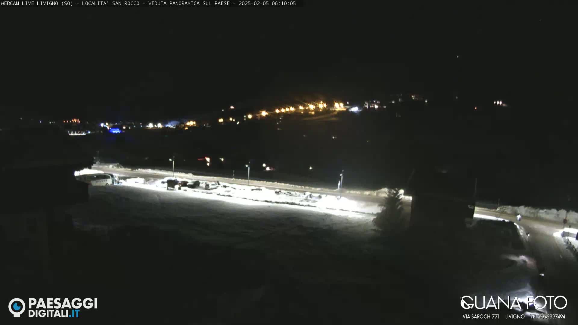 A nighttime, snow-covered landscape is illuminated by artificial lights, showing a road, parked vehicles, and a distant village glowing in the background.