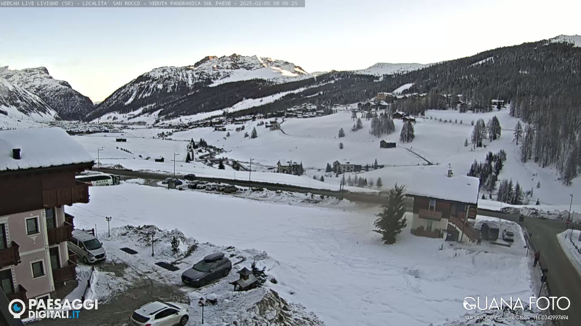 A snow-covered mountain valley village is seen under a clear, bright sky, with several buildings and parked cars visible in the foreground.