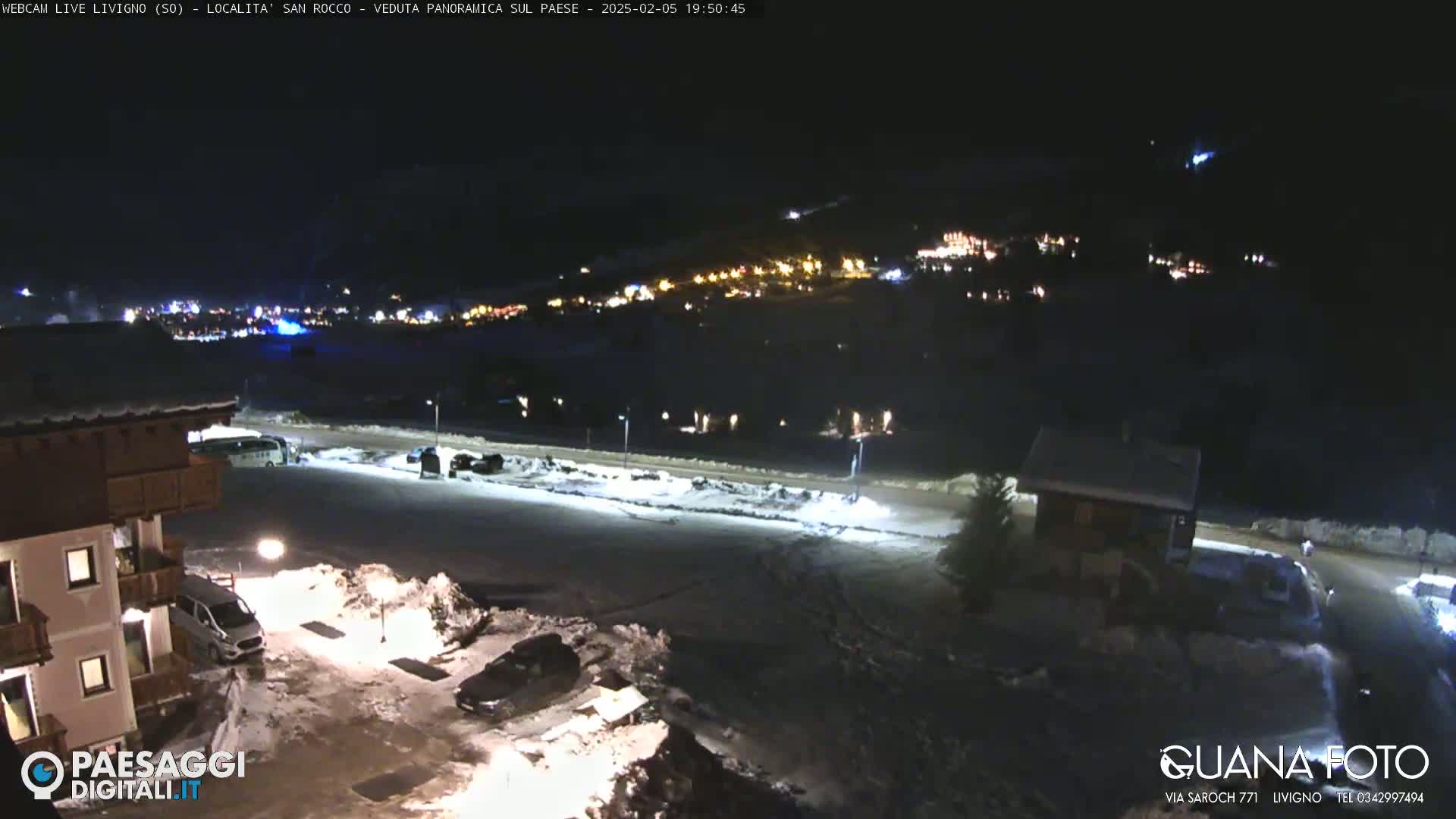 A nighttime view of a snow-covered parking lot and buildings, with a distant village illuminated against a dark sky.