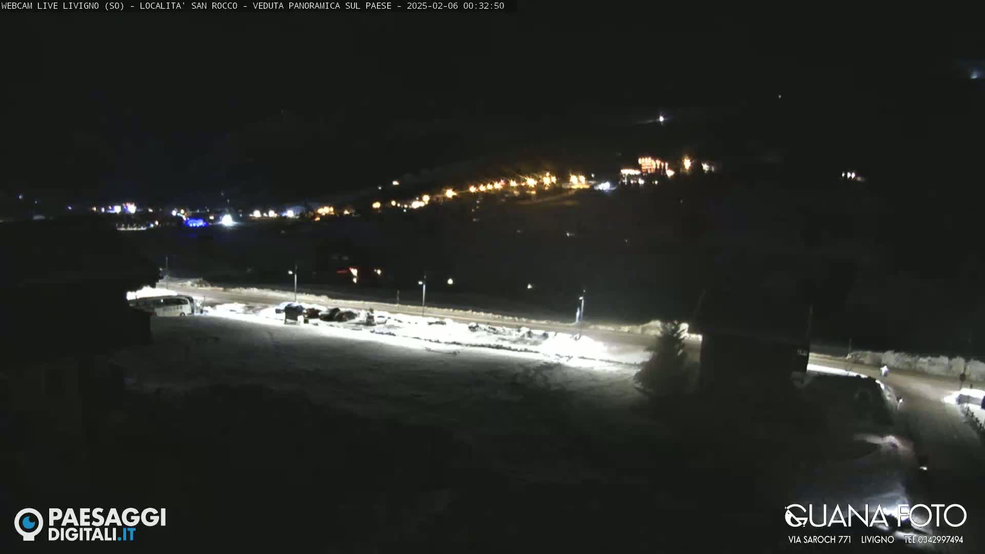 A nighttime, snowy scene shows a village illuminated with lights, a road, and several parked vehicles.