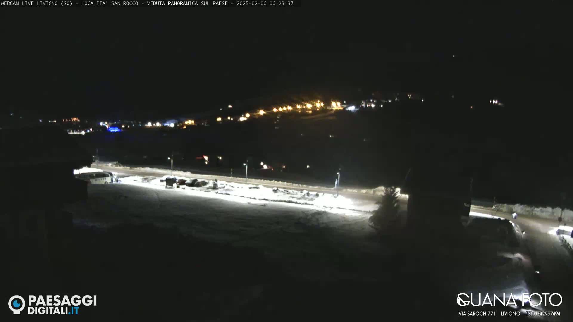 A nighttime, snow-covered parking lot and road are illuminated, with a village of lights visible in the distance.